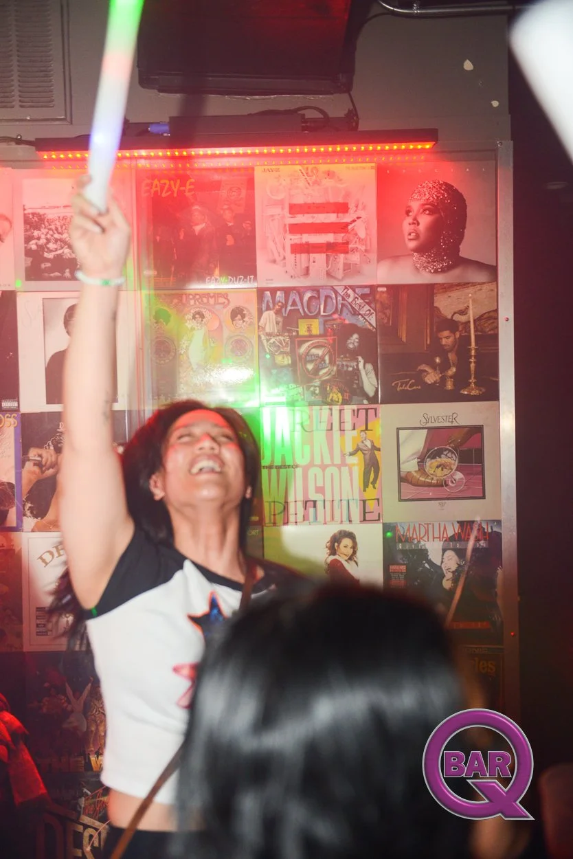 Person smiling and raising an arm in a lively bar or nightclub with a wall covered in music posters and album covers in the background.