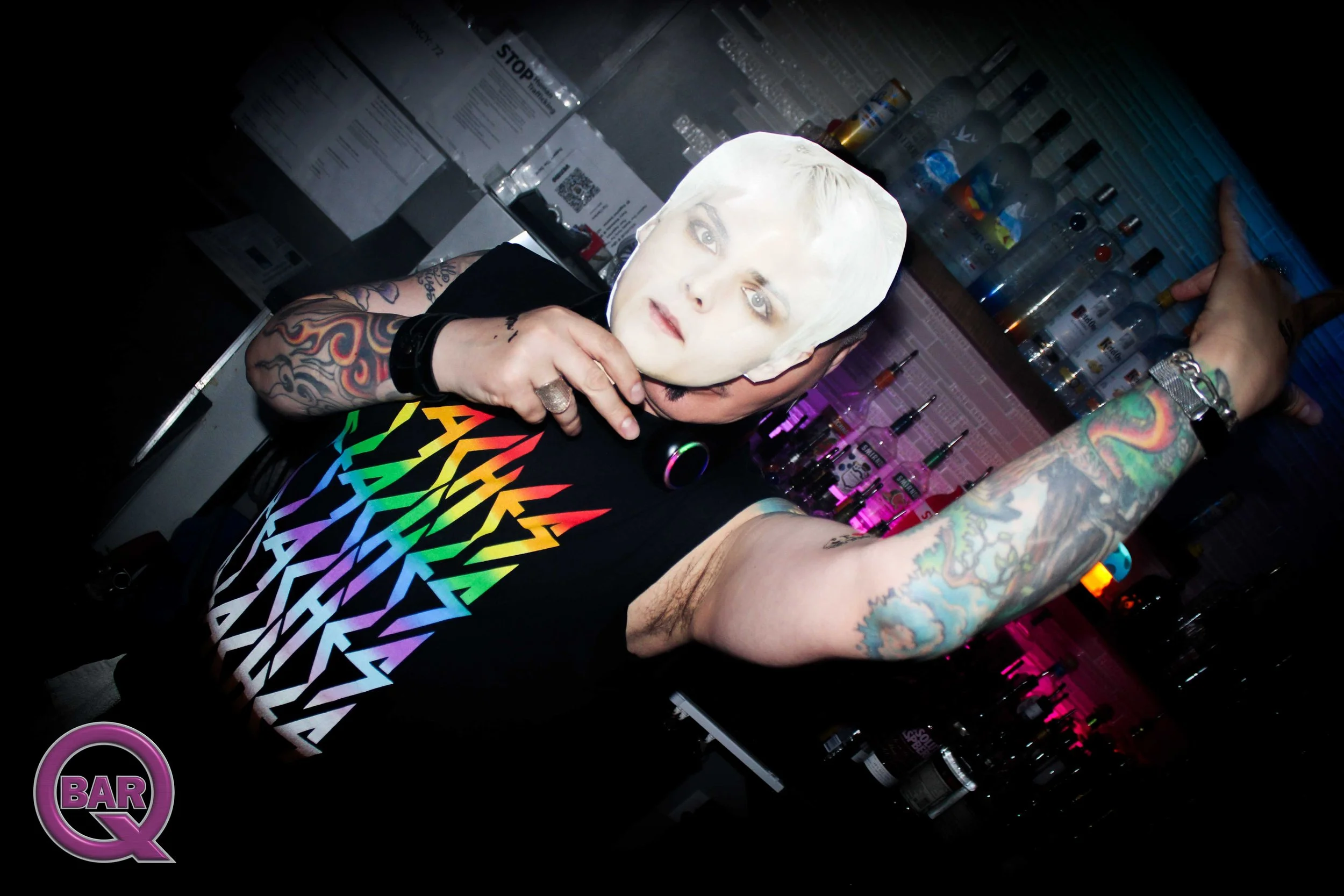 Person with tattoos on their arms wearing a rainbow-colored shirt, holding a mask of a woman's face, behind a bar with various bottles and items.