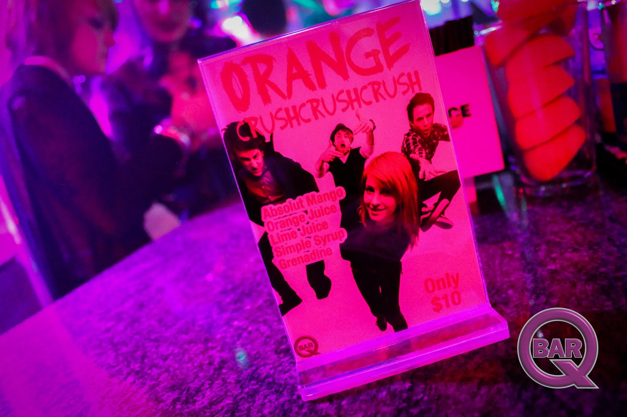 Pink promotional sign for orange crush drinks with images of two women and one man making expressive gestures. The sign lists flavors, including mango, orange juice, lime juice, simple syrup, and grenadine. The price is $10, and the background has pu
