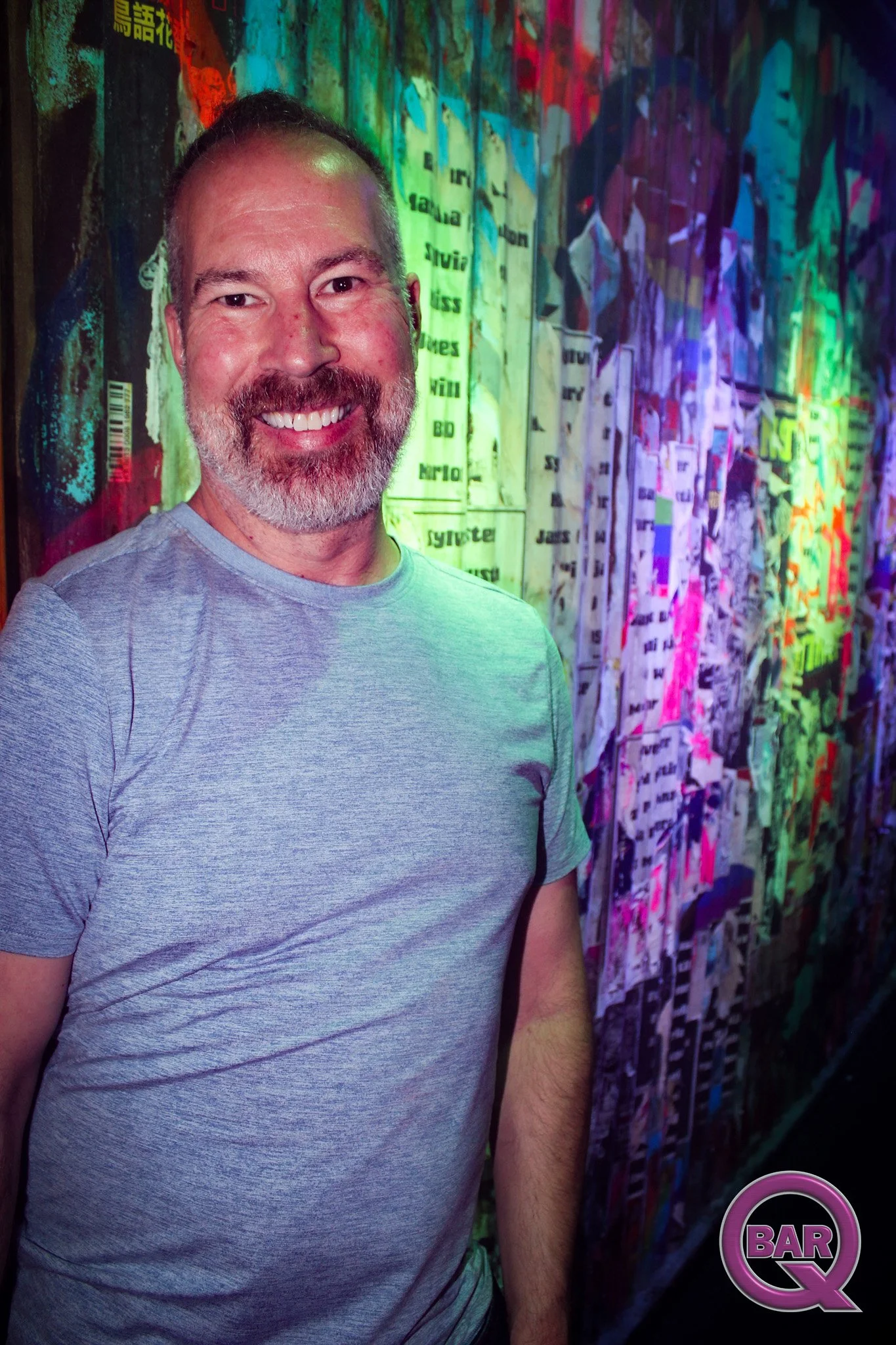 A smiling man with a beard and short hair wearing a light gray t-shirt, standing in front of a colorful, abstract background with a mix of vibrant colors and patterns.