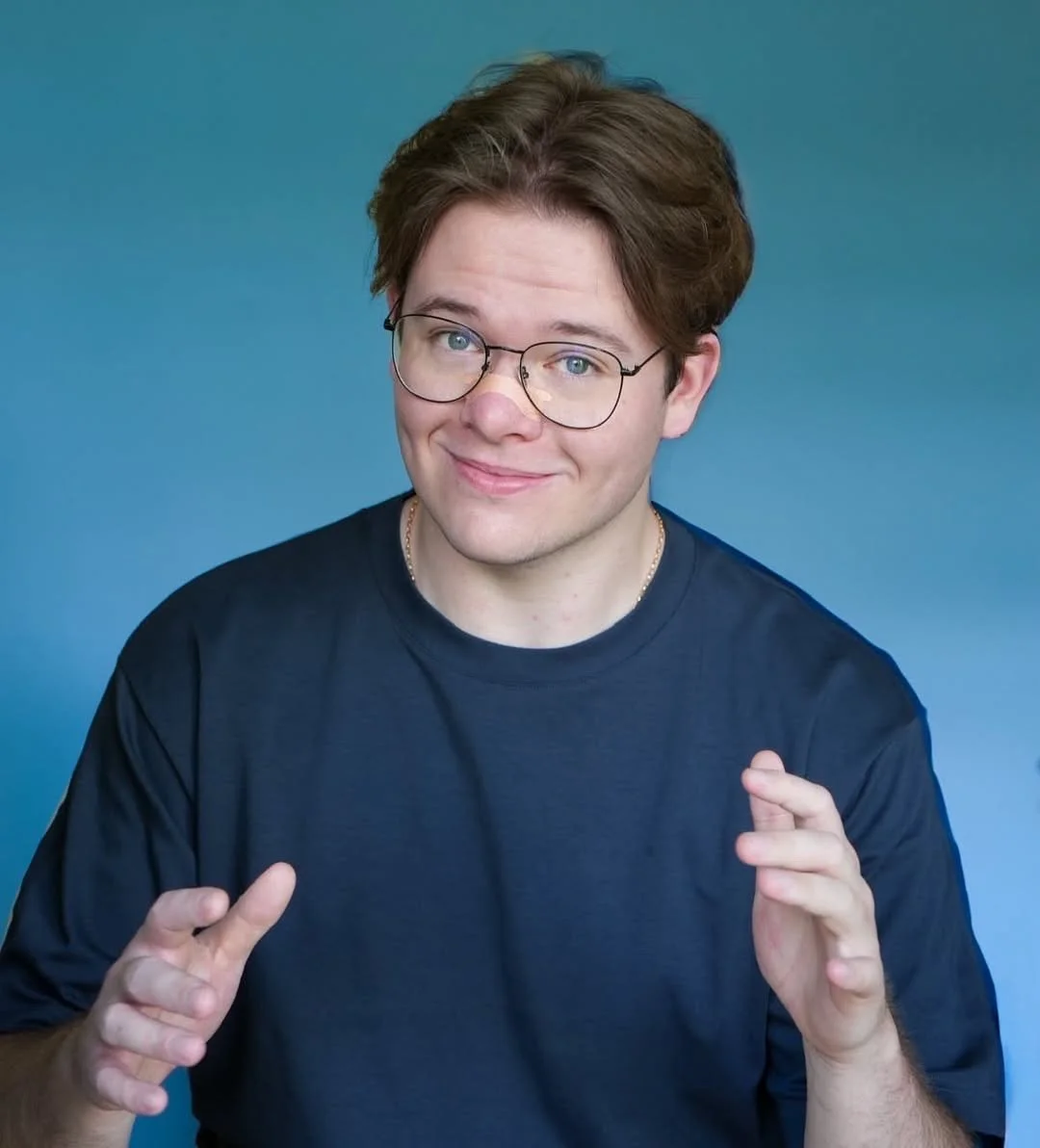 A young man with glasses, smiling and making a playful gesture with his hands against a blue background.
