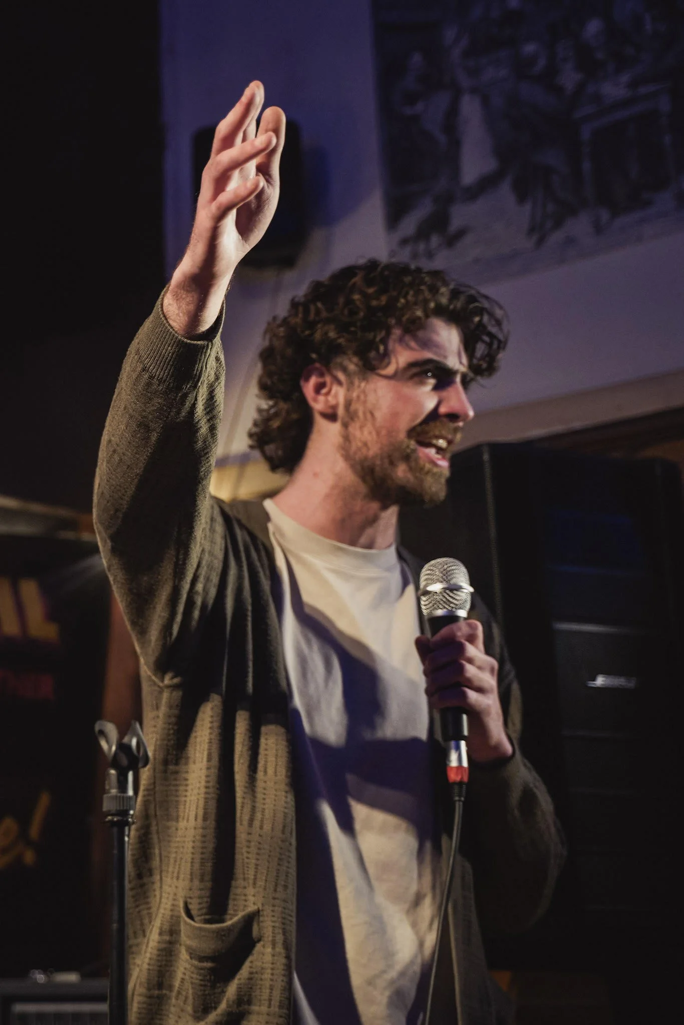 A man with curly hair and a beard is holding a microphone and raising his right hand, speaking or singing on stage with a dark background and some musical equipment.
