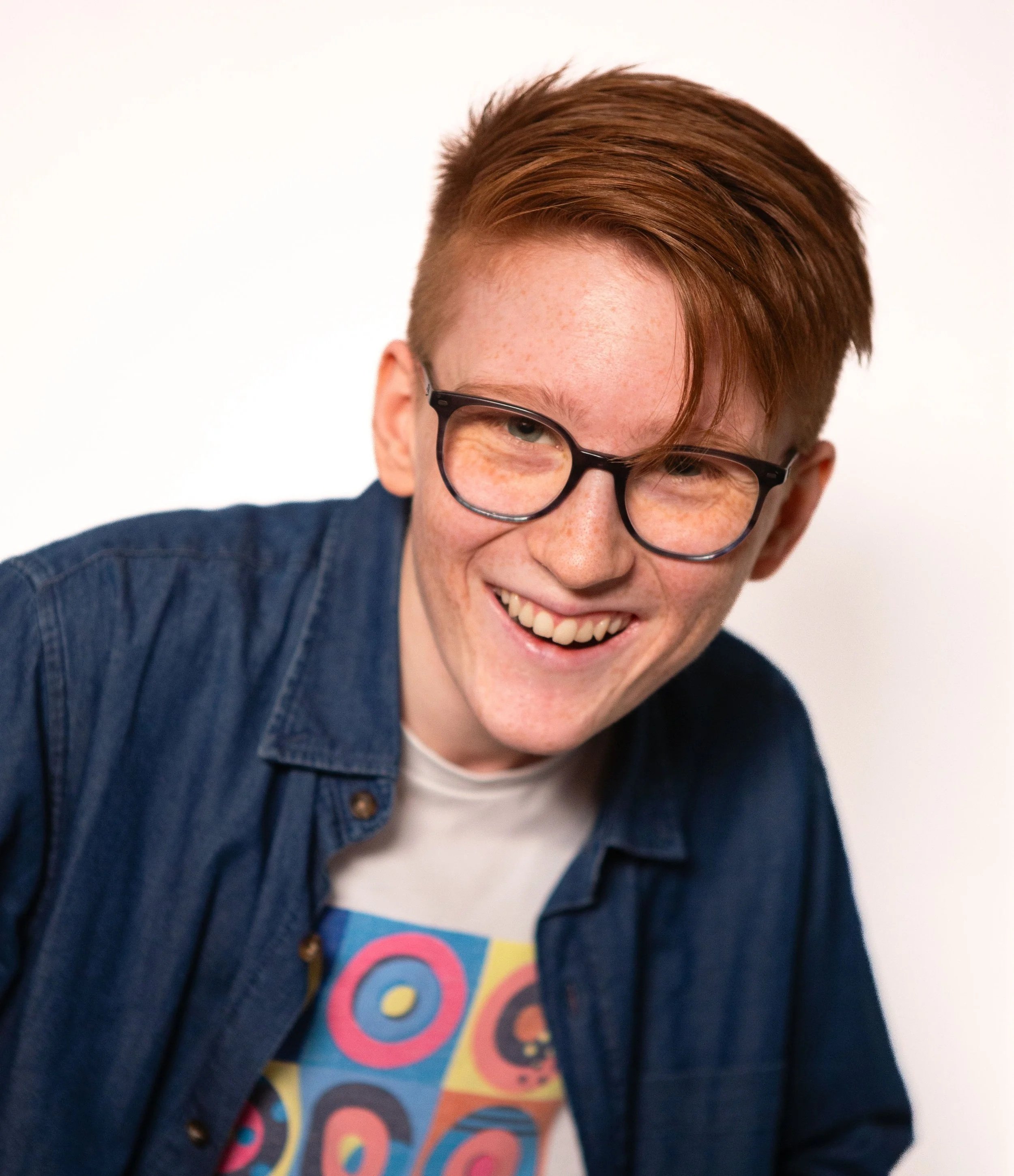 A person with light skin and reddish hair, partially visible from the nose up, wearing black glasses. They are smiling and wearing a denim jacket over a colorful shirt with circular patterns.