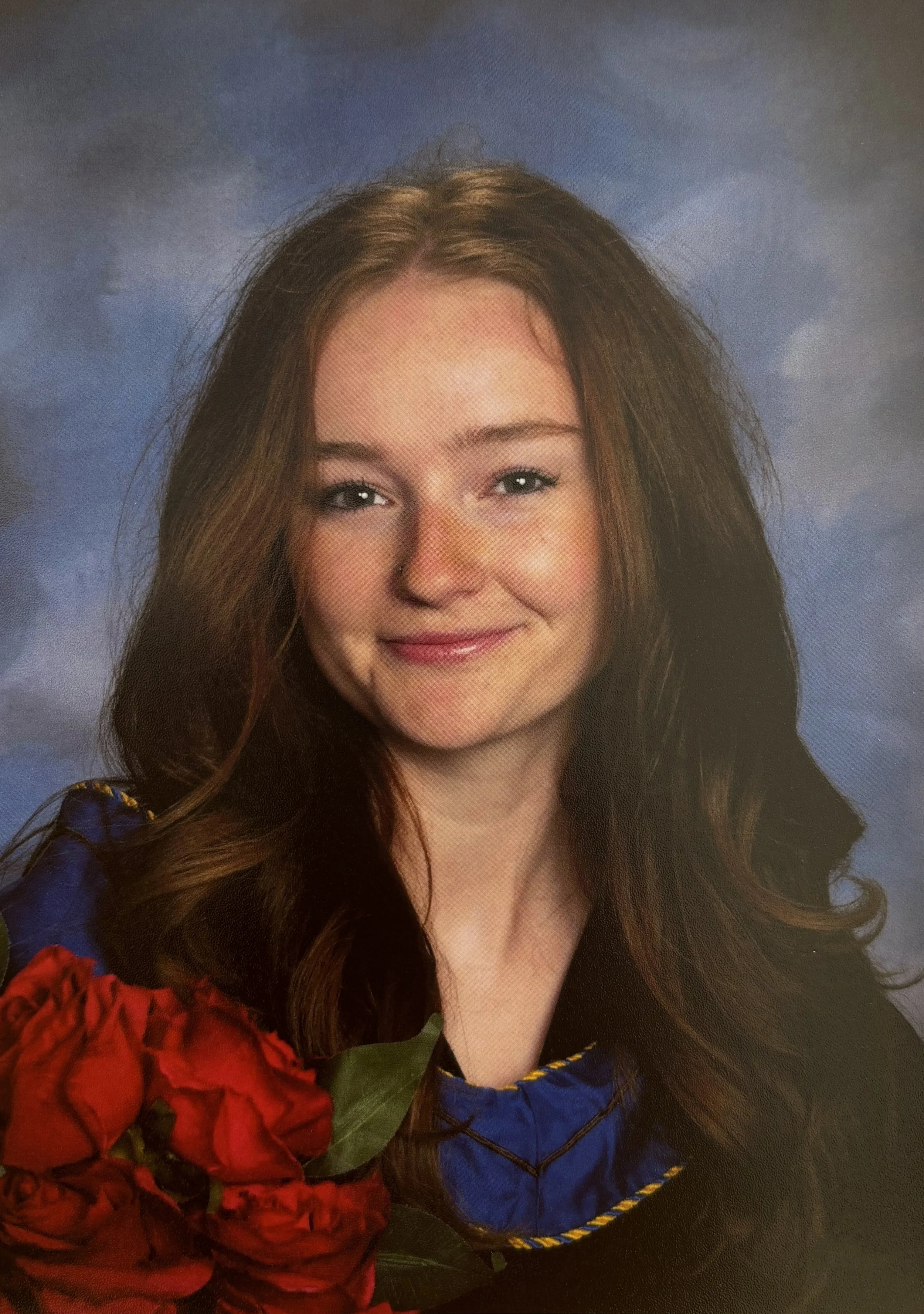 A portrait of a young woman with long, wavy brown hair and a light complexion, smiling softly. She is wearing a colorful dress with blue, red, and yellow details and is holding a large bouquet of red flowers. The background is a cloudy blue sky.