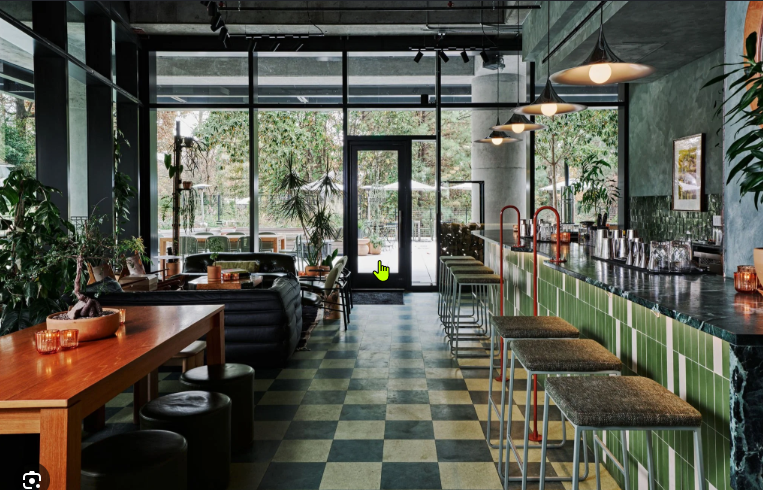Interior of a modern cafe with green tiled bar, bar stools, black leather seating, large windows, and plants.