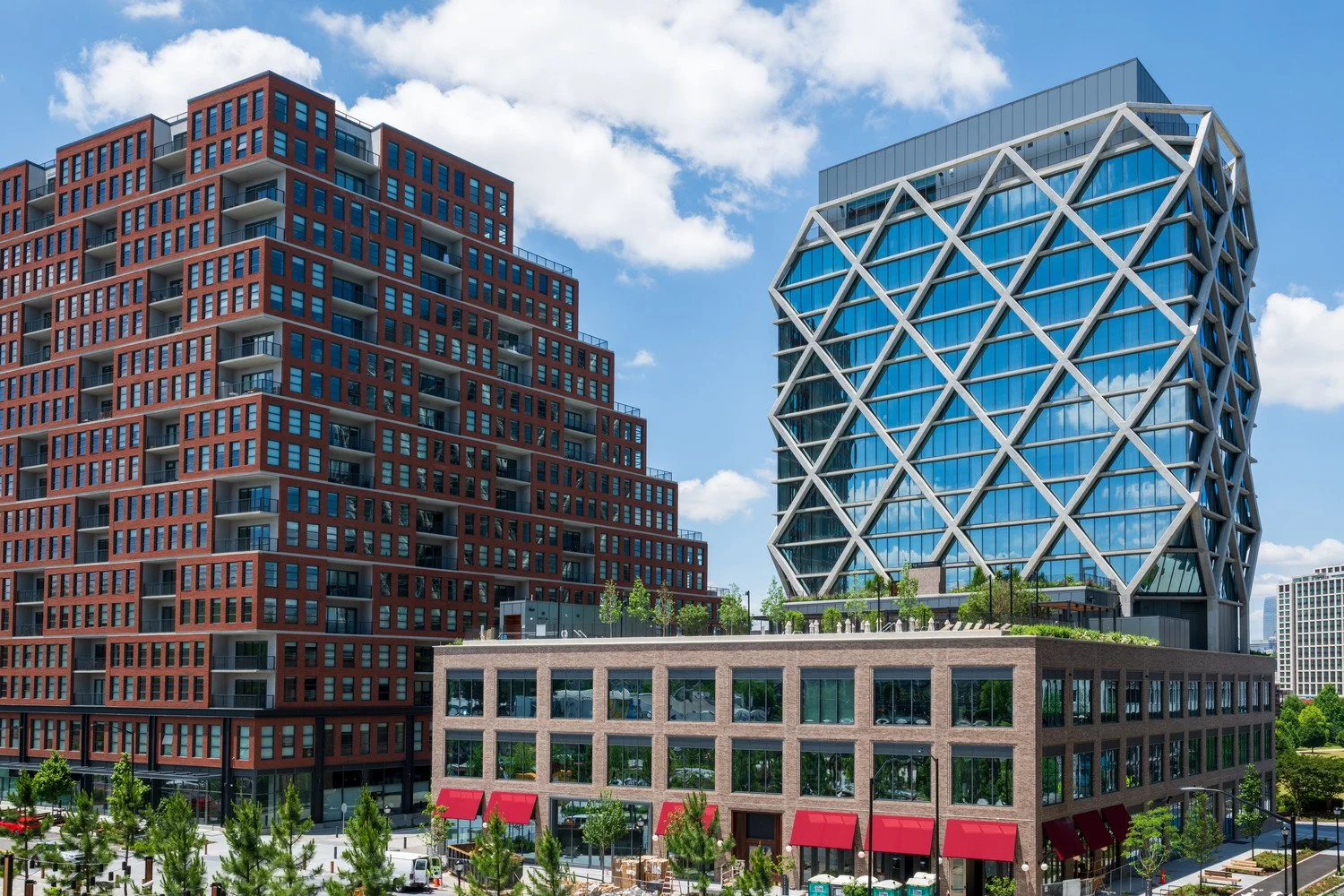 Modern city buildings with a clear blue sky and clouds, including a red-brick apartment building and a glass office tower with a unique geometric design.