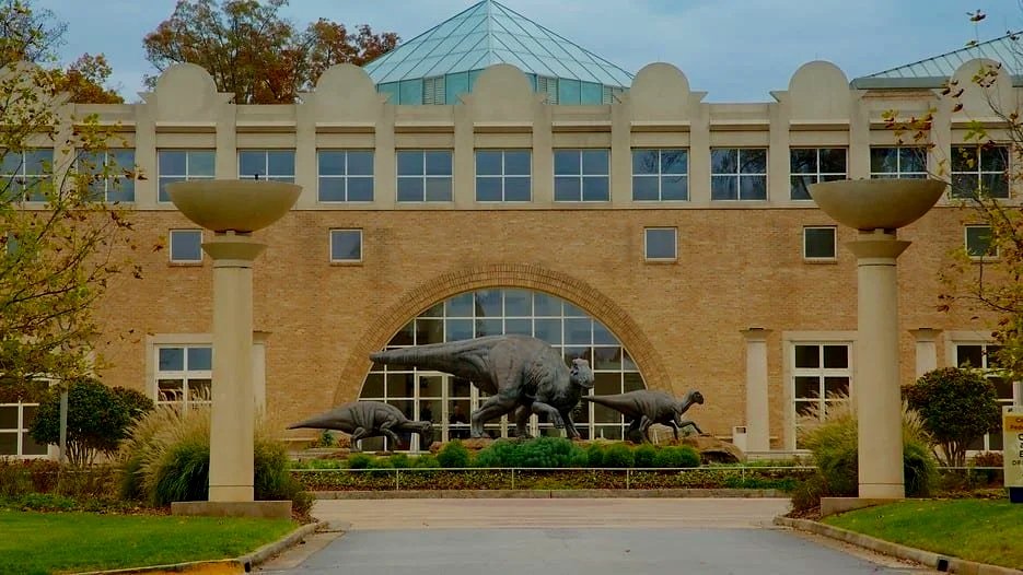 A building with a modern, glass pyramid roof, brick facade, and dinosaur sculptures in front, surrounded by landscaped greenery and tall lamp posts.