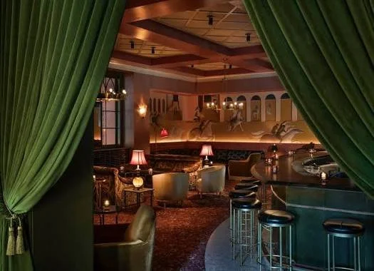 Interior of a lounge or bar with green curtains framing the view, showing seating areas with chairs and tables, a bar counter with stools, and warm lighting.