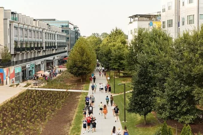 Urban park with people walking along a paved path, surrounded by trees, apartment buildings, and outdoor seating areas on a sunny day.
