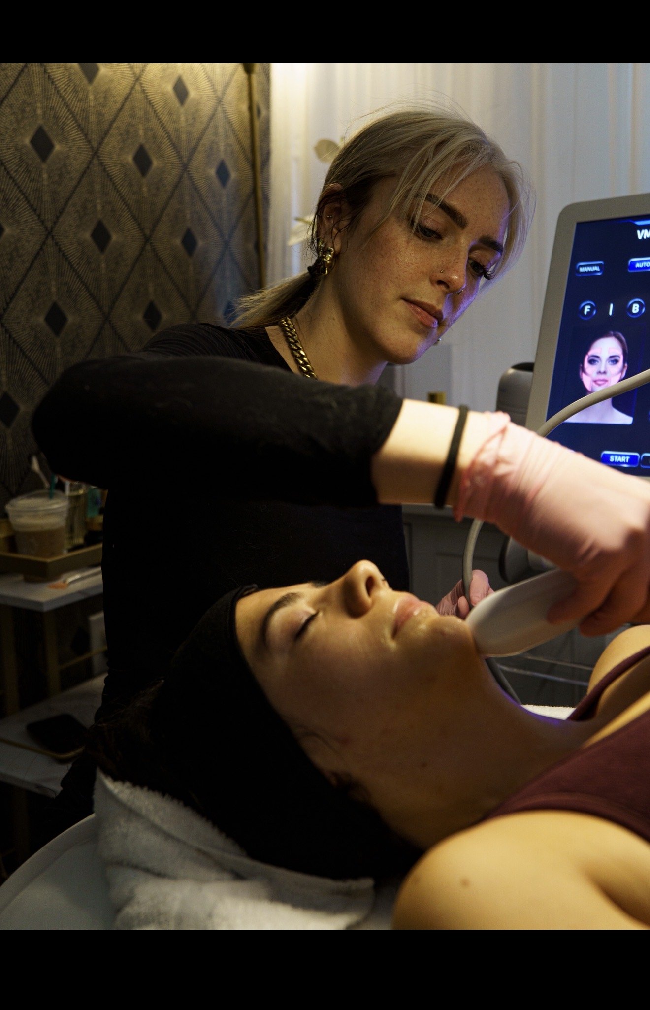 A美容師 performing a facial treatment on a reclining woman in a spa or clinic, with a monitor displaying a digital face in the background.