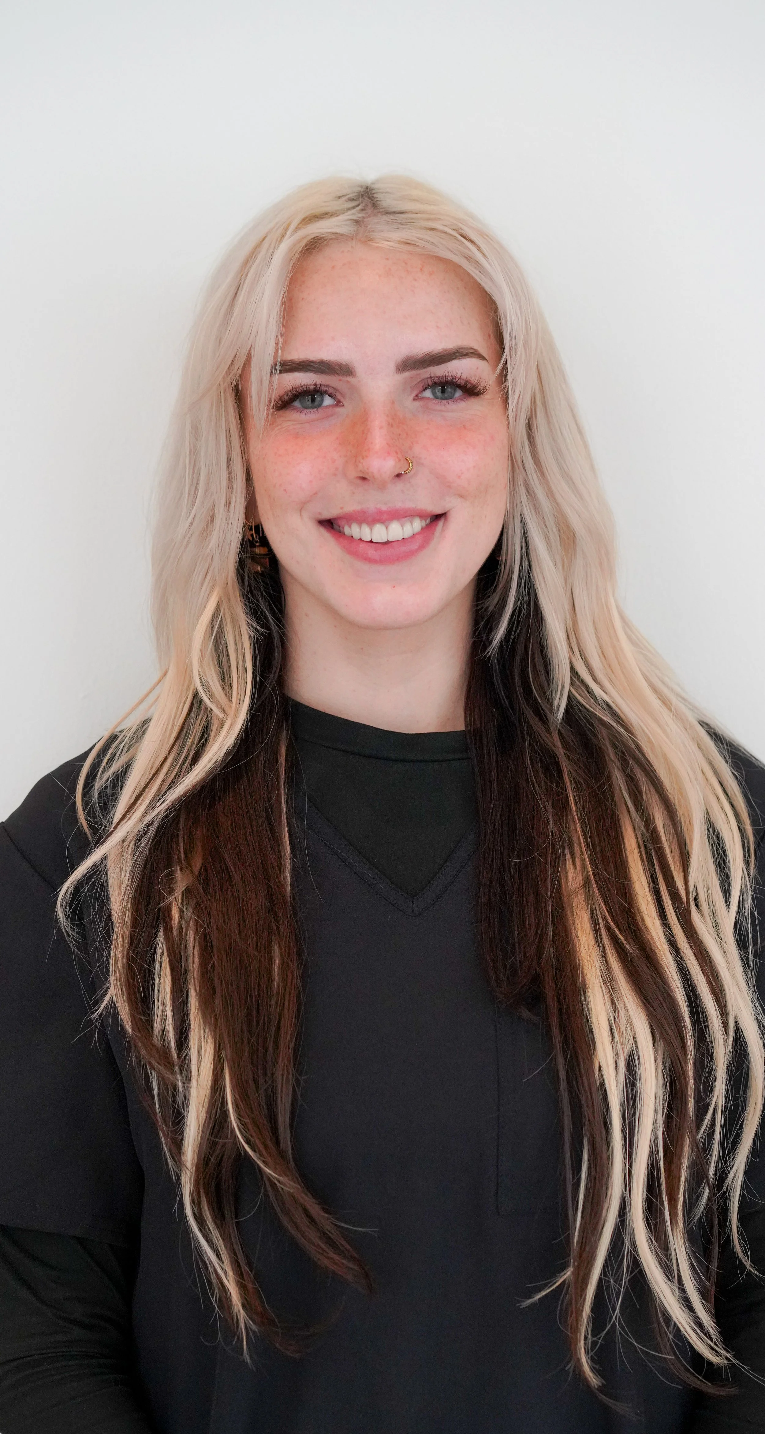 A young woman with long blonde hair and light blue eyes smiling, wearing a black top against a plain white background.