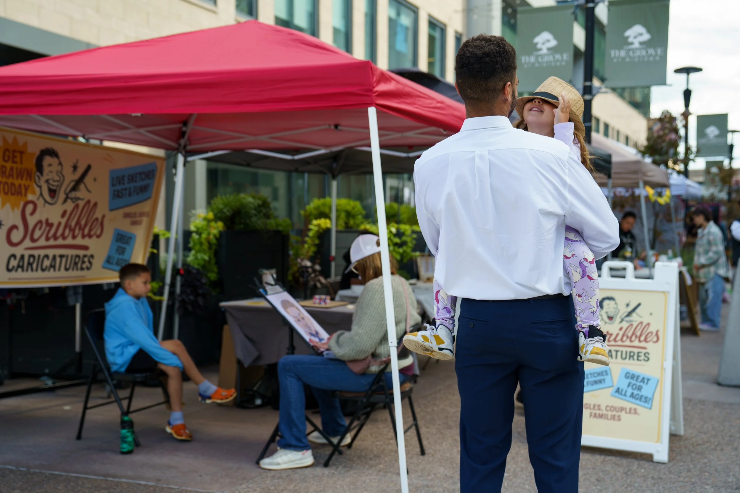 Jazmyne Throumoulos wears her dad Nik’s hat while her brother poses for a caricature drawing at The Lucky Flea in downtown Rochester on Sept. 7, 2025 in Rochester, N.Y. Nik took his son and daughter to The Lucky Flea to explore the city, and after, t