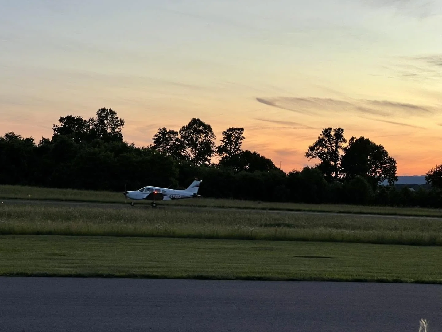 Small private airplane on a runway at sunset, surrounded by trees, with a colorful sky overhead.
