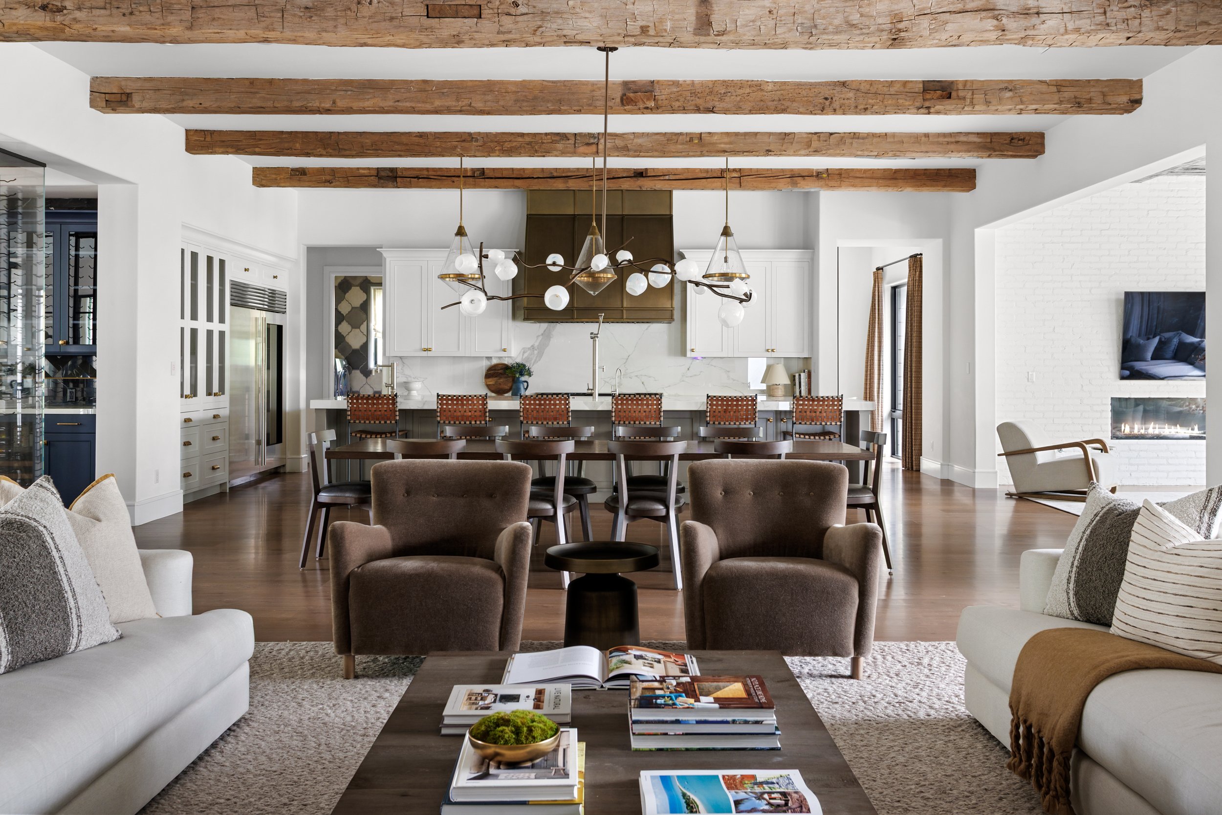 Living room with white sofas, a coffee table with books and magazines, and beige armchairs, leading to an open kitchen with white cabinets, a marble backsplash, and wooden beams on the ceiling.
