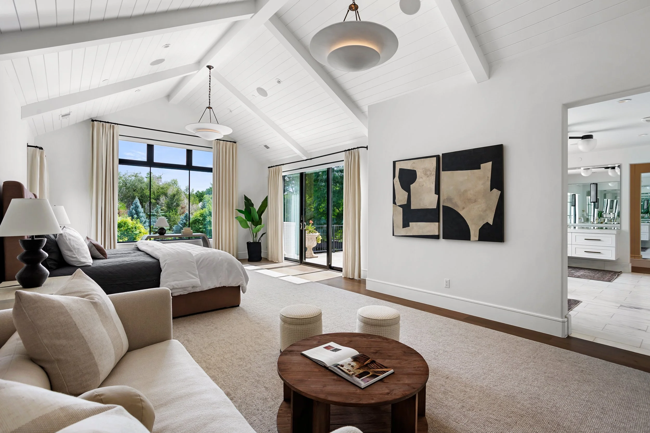 Bright living room with white walls, vaulted ceiling, modern pendant lights, large windows with curtains, a bed, armchairs, potted plant, abstract wall art, and a view of lush greenery outside