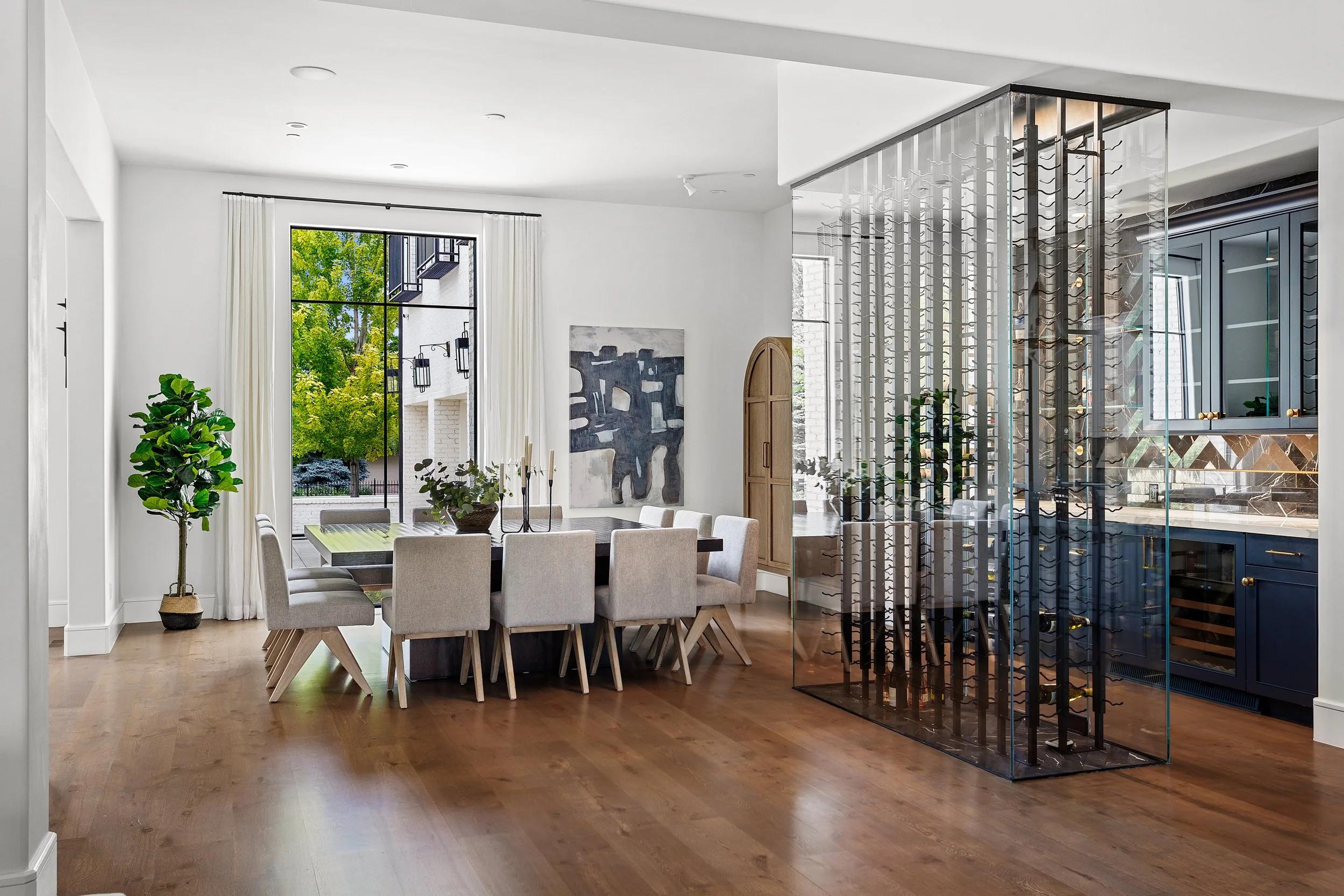 Modern dining room with large window, white walls, wooden floor, and a six-seater dining table with beige chairs. There is a green potted plant and a piece of abstract art on the wall. A glass wine storage unit with a wine rack and cabinetry is visib