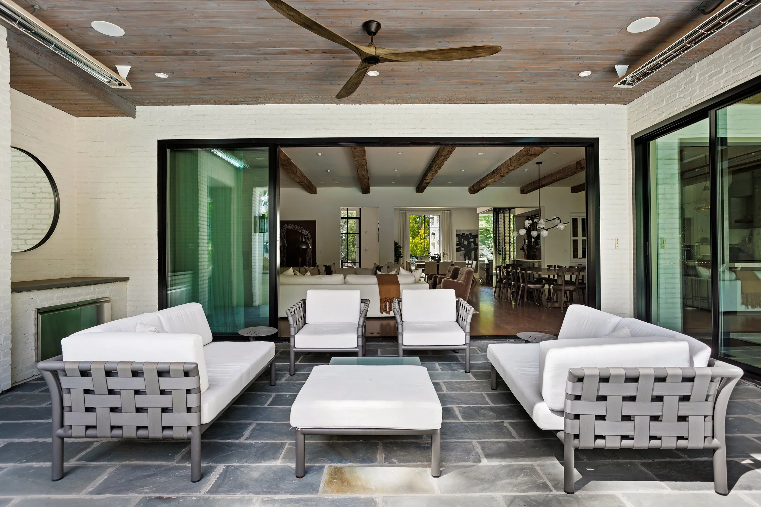 A modern indoor-outdoor living space with white brick walls, wooden ceiling with beams, ceiling fan, and large glass sliding doors. The outdoor area features white cushioned seating and a coffee table, with a stone floor.