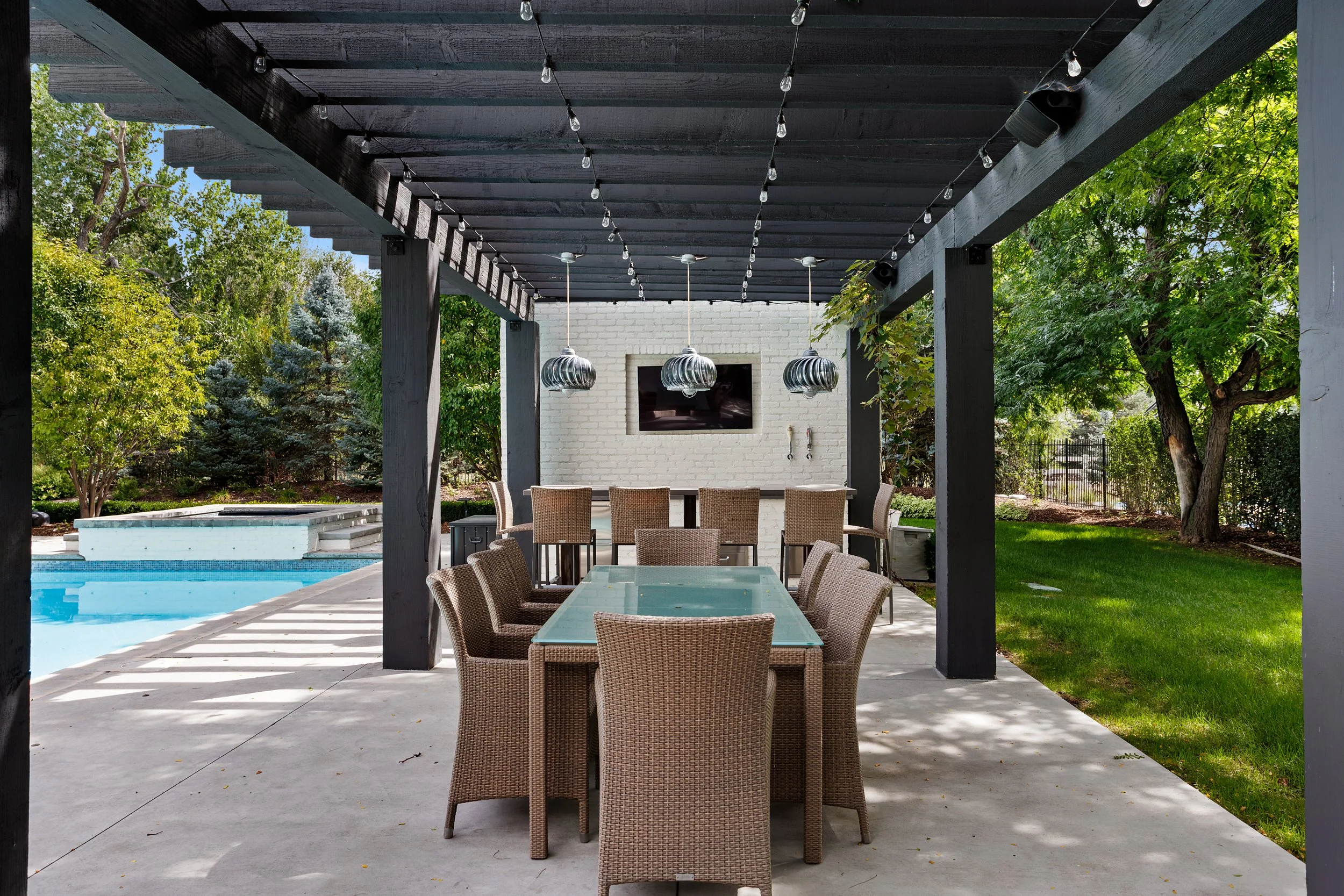 An outdoor patio with a dining area under a pergola, trees and a swimming pool in the background.