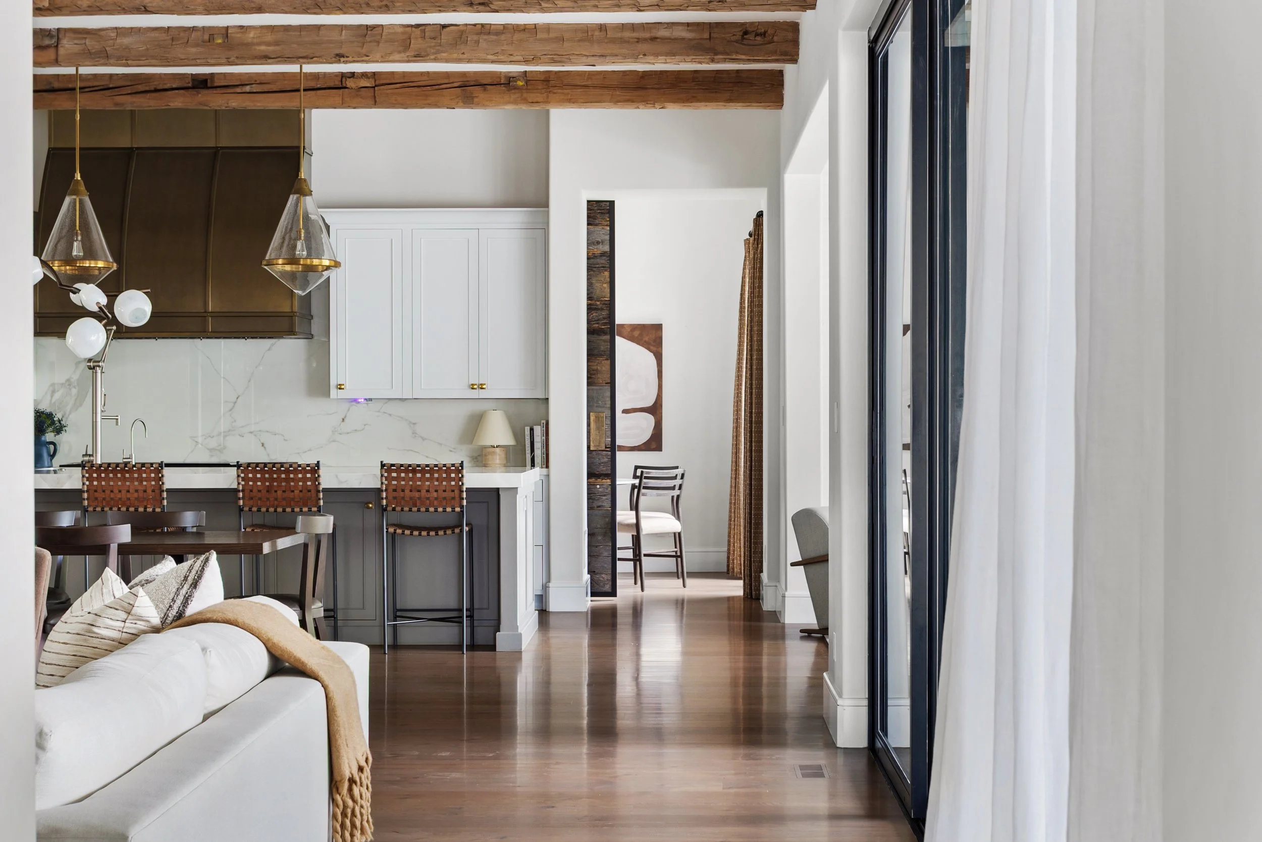 Open-concept living space with wooden beams, large windows with white curtains, a white sofa with pillows and a throw blanket, a dining area with chairs, and a kitchen with white cabinets and gold accents.
