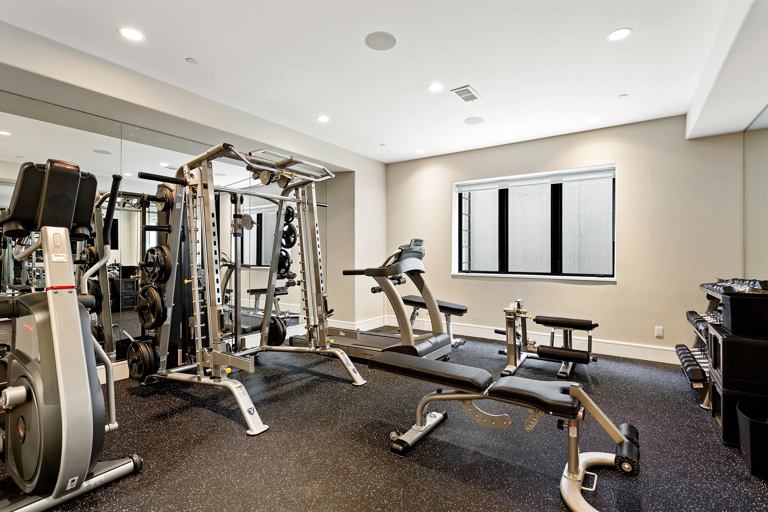 Indoor gym with various exercise equipment including treadmills, weight machines, and free weights, illuminated by ceiling lights and a window.