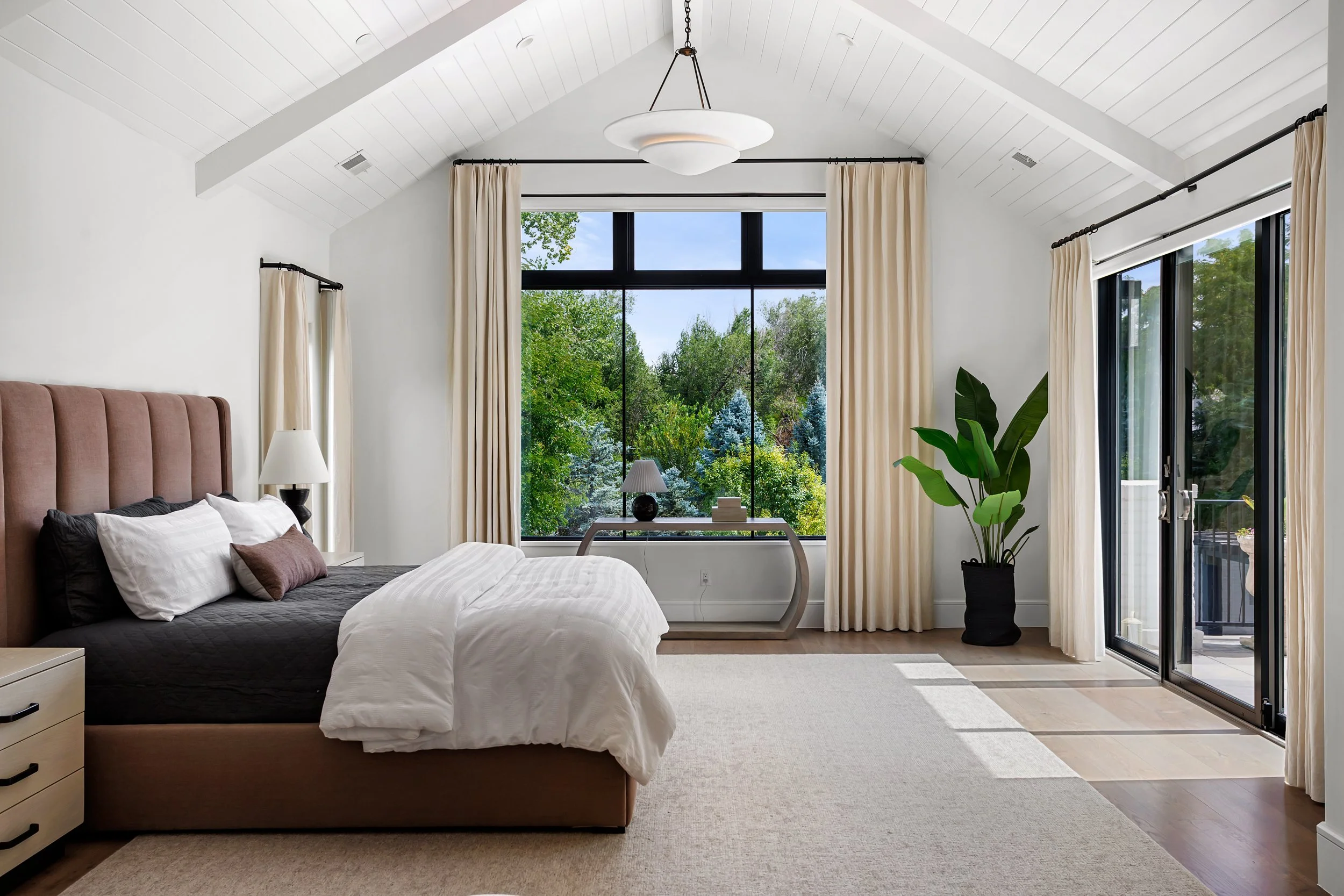 A bedroom with a large window showing trees outside, white walls, beige curtains, a bed with white and dark pillows, a nightstand, lamps, and a large potted plant.