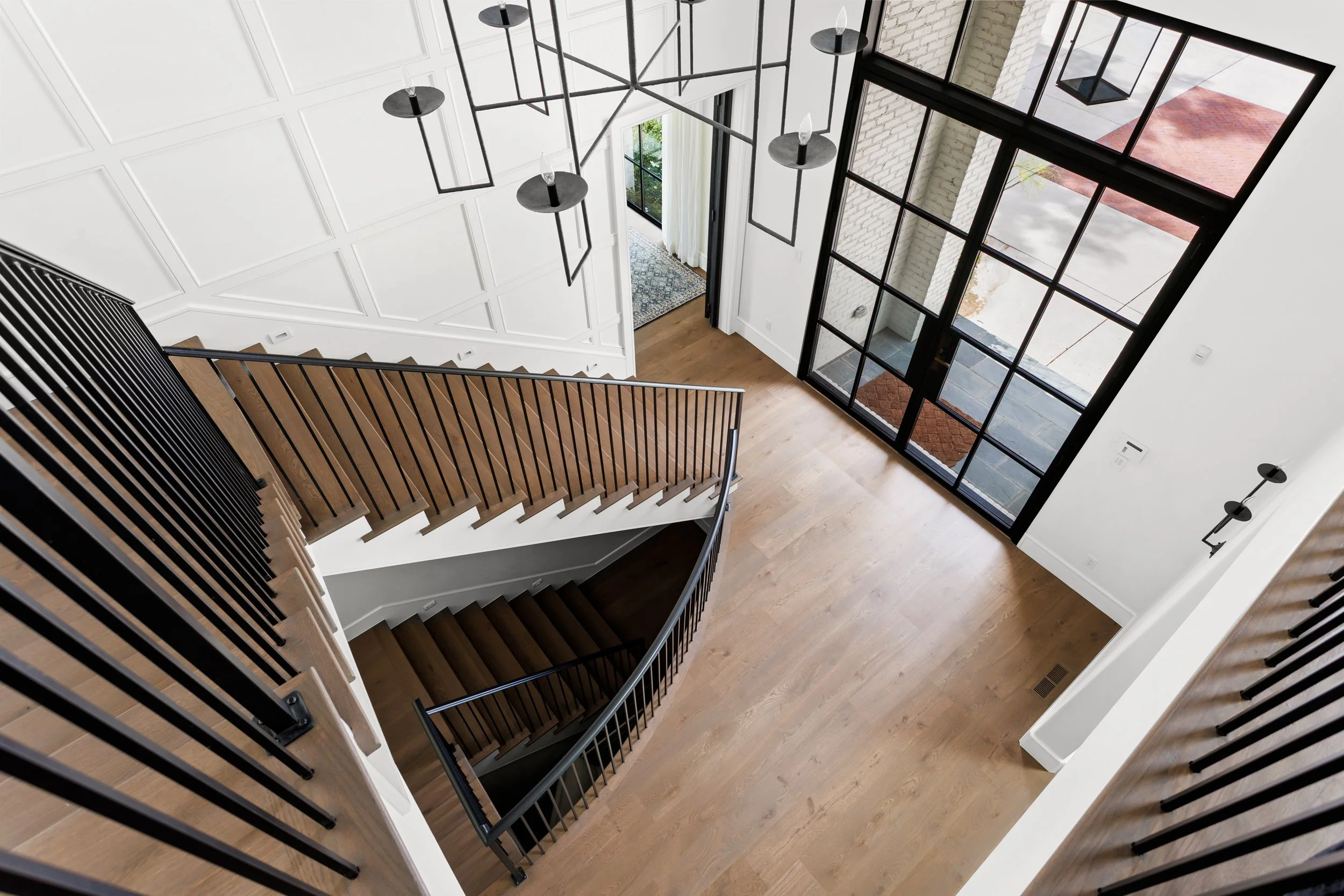 Interior view of a modern staircase overlooking a foyer with large glass windows, white walls, and wooden flooring.