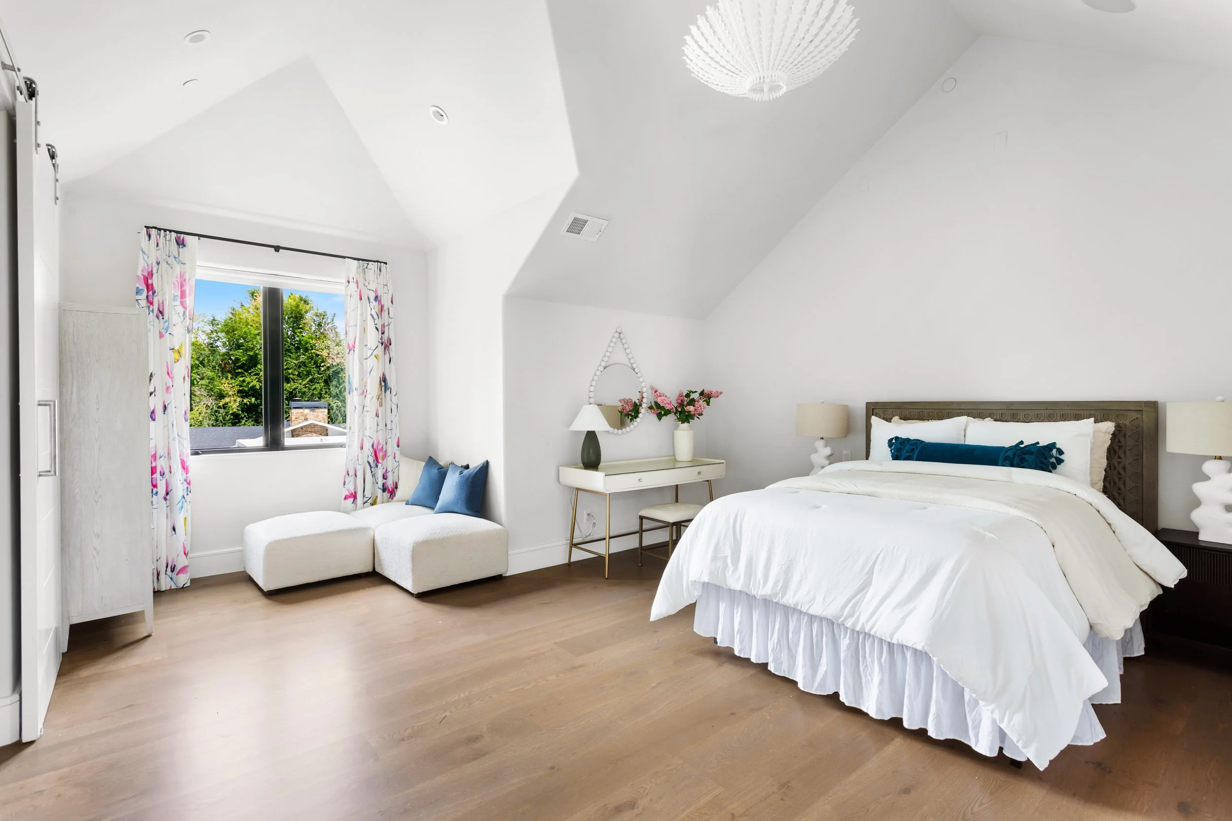 Bright bedroom with a white bed, golden accents, a white vanity with pink flowers, a beige chair with blue pillows, a window with floral curtains, and wooden flooring.