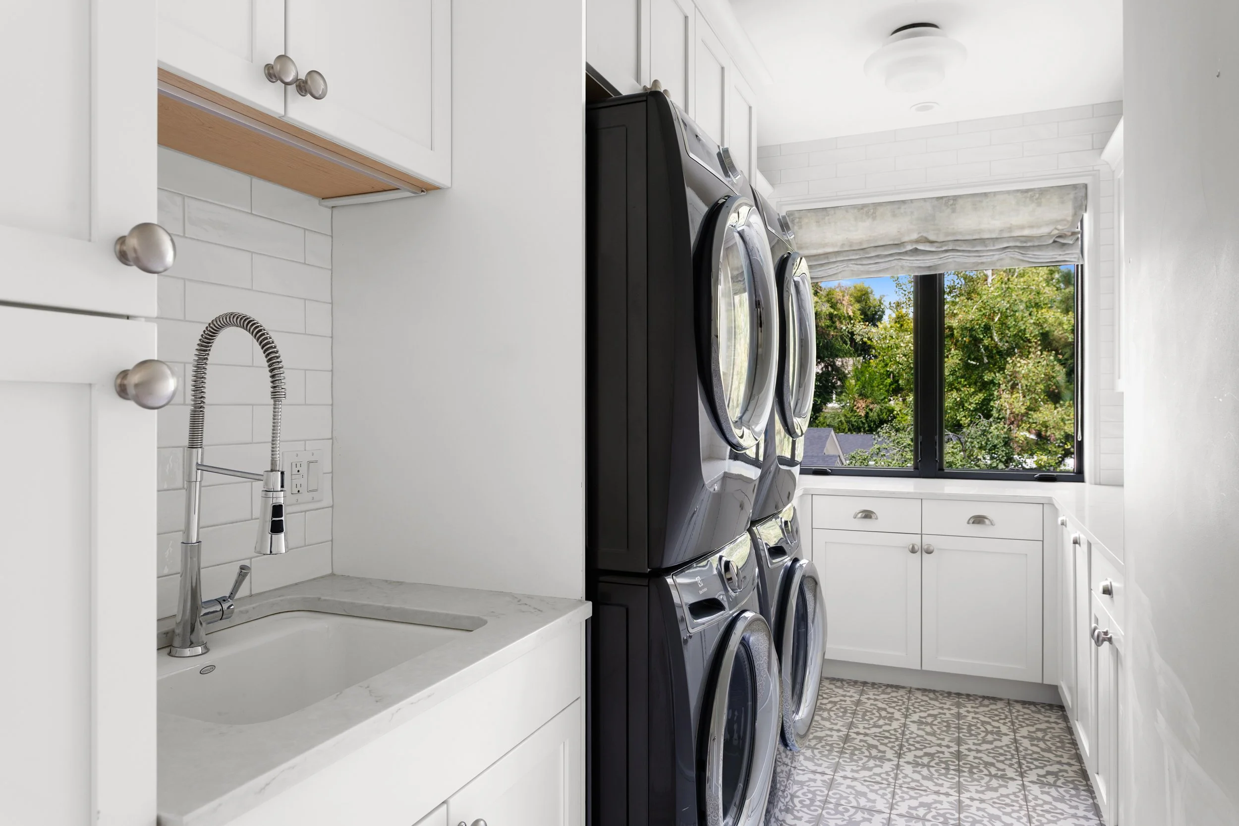 A laundry room with black washer and dryer units, white cabinets, a white marble countertop with a sink, and a window showing trees outside.
