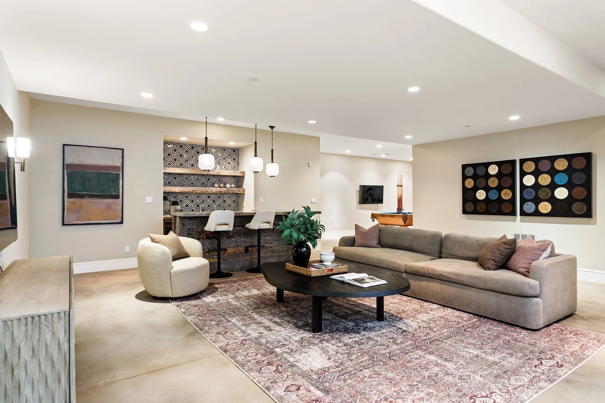 Living room with beige sofa, patterned area rug, round black coffee table, white armchair, decorative wall art, a plant, and a bar area with hanging pendant lights.