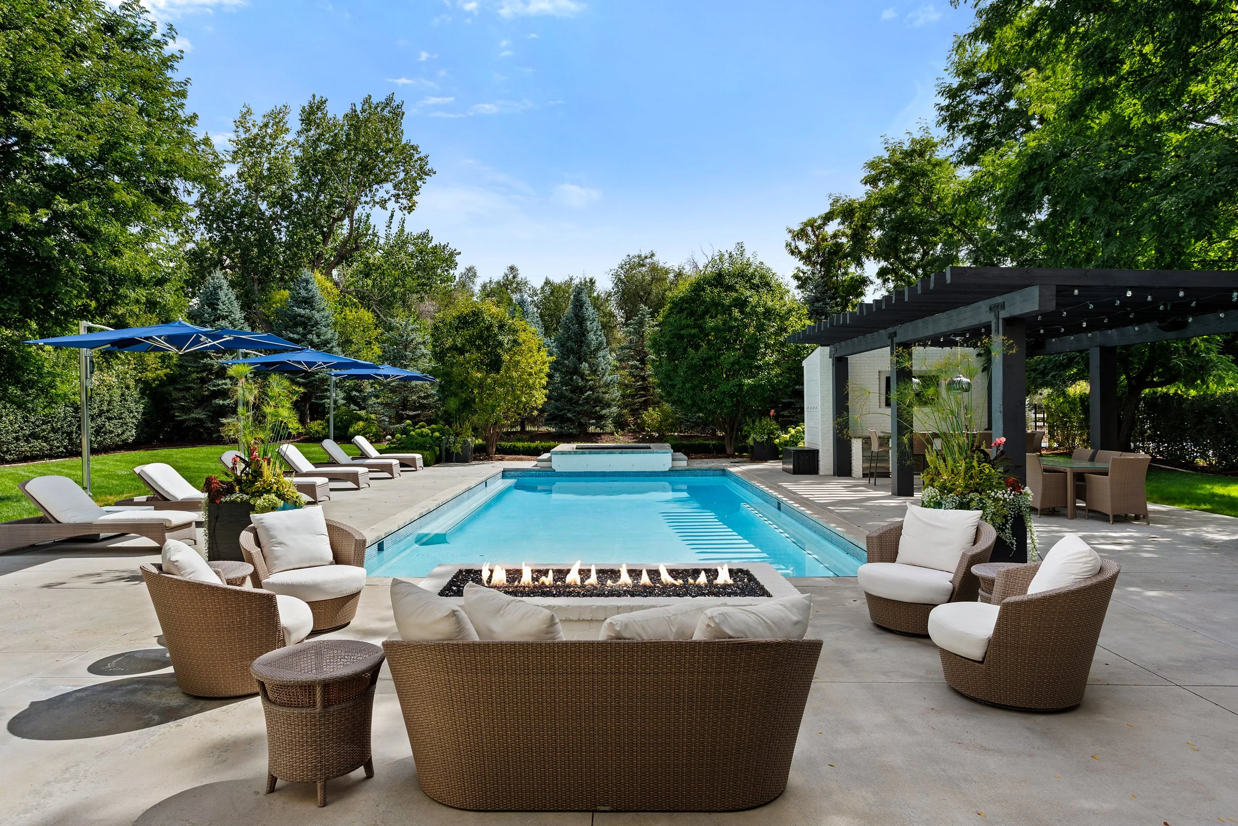 A backyard swimming pool area with lounge chairs, a fire pit, and umbrellas, surrounded by lush green trees and a sitting area under a pergola with string lights.
