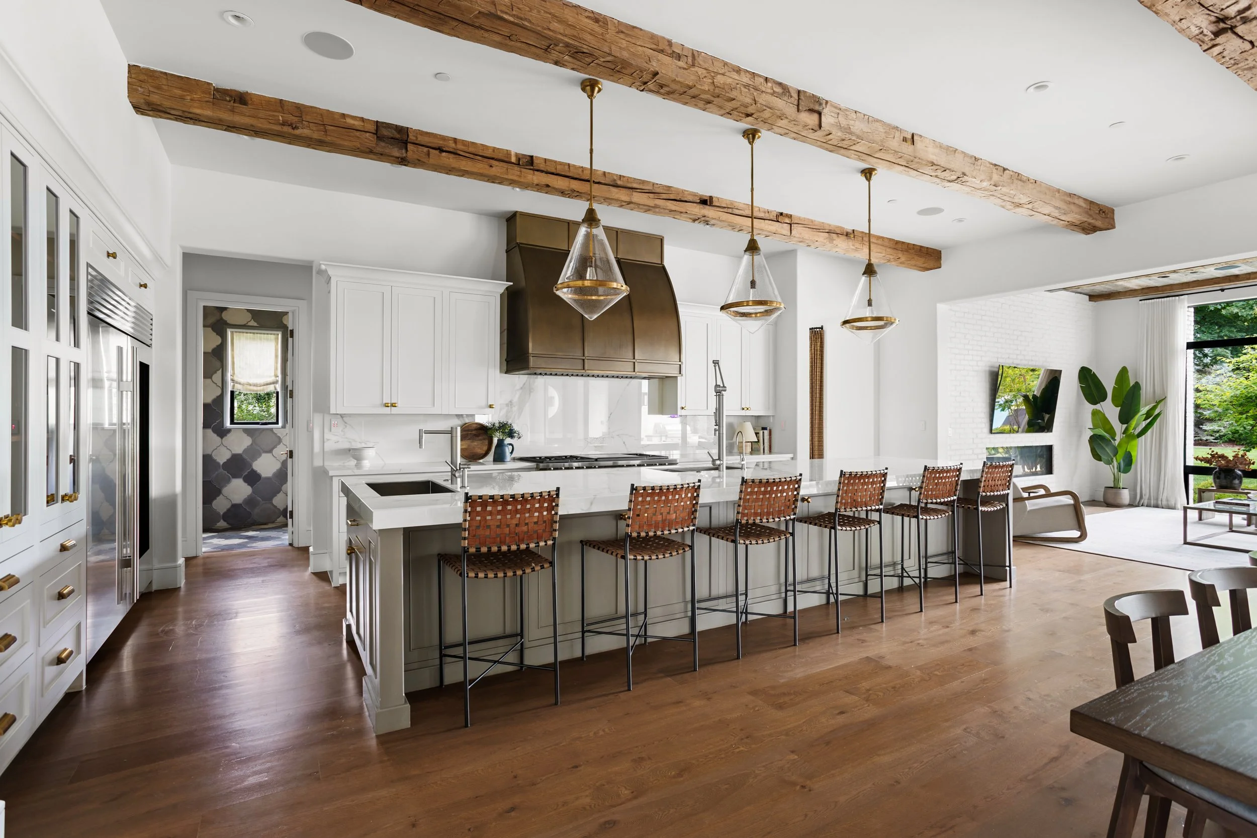 A modern kitchen and living room with white cabinets, a large white island with a marble countertop, wooden bar stools, a stainless steel range hood, and pendant lights hanging from wooden beams on the ceiling. There are large windows with views of g
