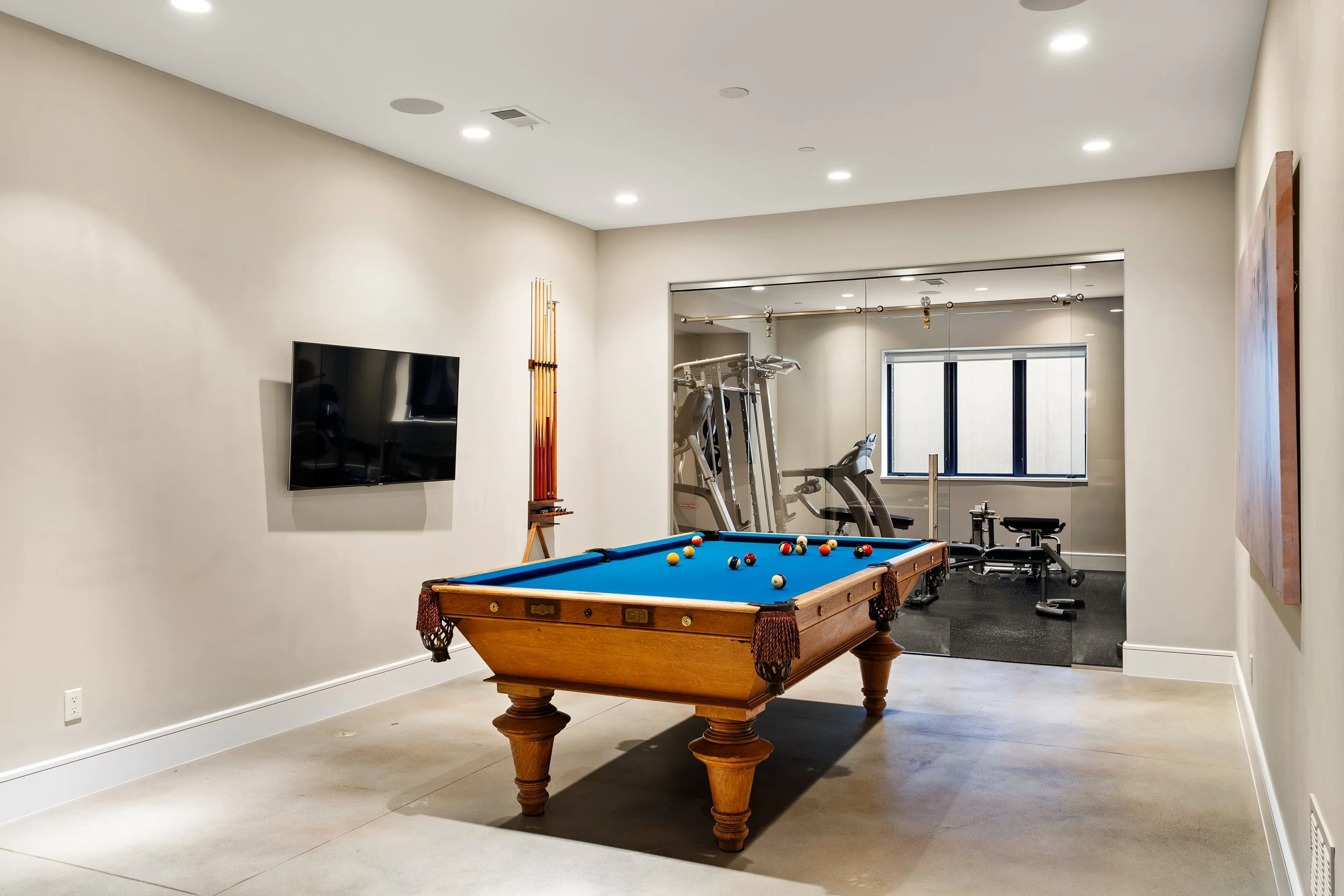 A recreational room featuring a blue pool table with various pool balls on it. In the background, gym equipment including a treadmill and elliptical machine are visible, with a mirror reflecting the room and a window with a blind. The room has white 
