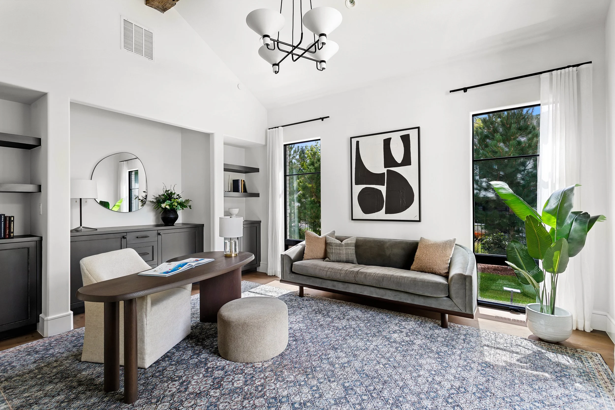 A modern living room with white walls, large windows with white curtains, a gray sofa with cushions, a round wooden coffee table, and a beige armchair with a matching ottoman. Decor includes a black sideboard with a flower vase, a mirror, and a black and white abstract art piece. There is a patterned area rug and a large potted plant near the windows. A black chandelier hangs from the ceiling.