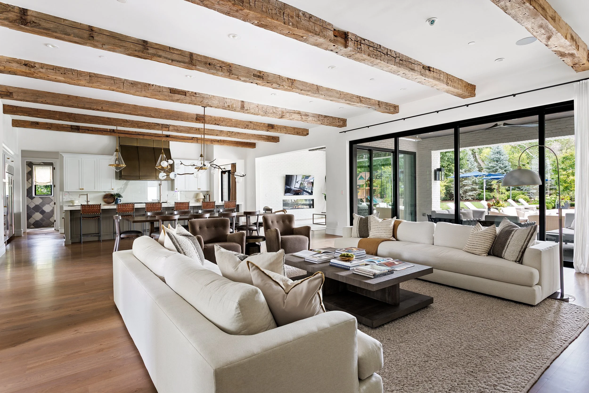 Open-concept living room and kitchen with exposed wooden beams on the ceiling, large windows and sliding glass doors, white couches and brown chairs, modern pendant lights, and a view of a backyard patio with lounge chairs and greenery.