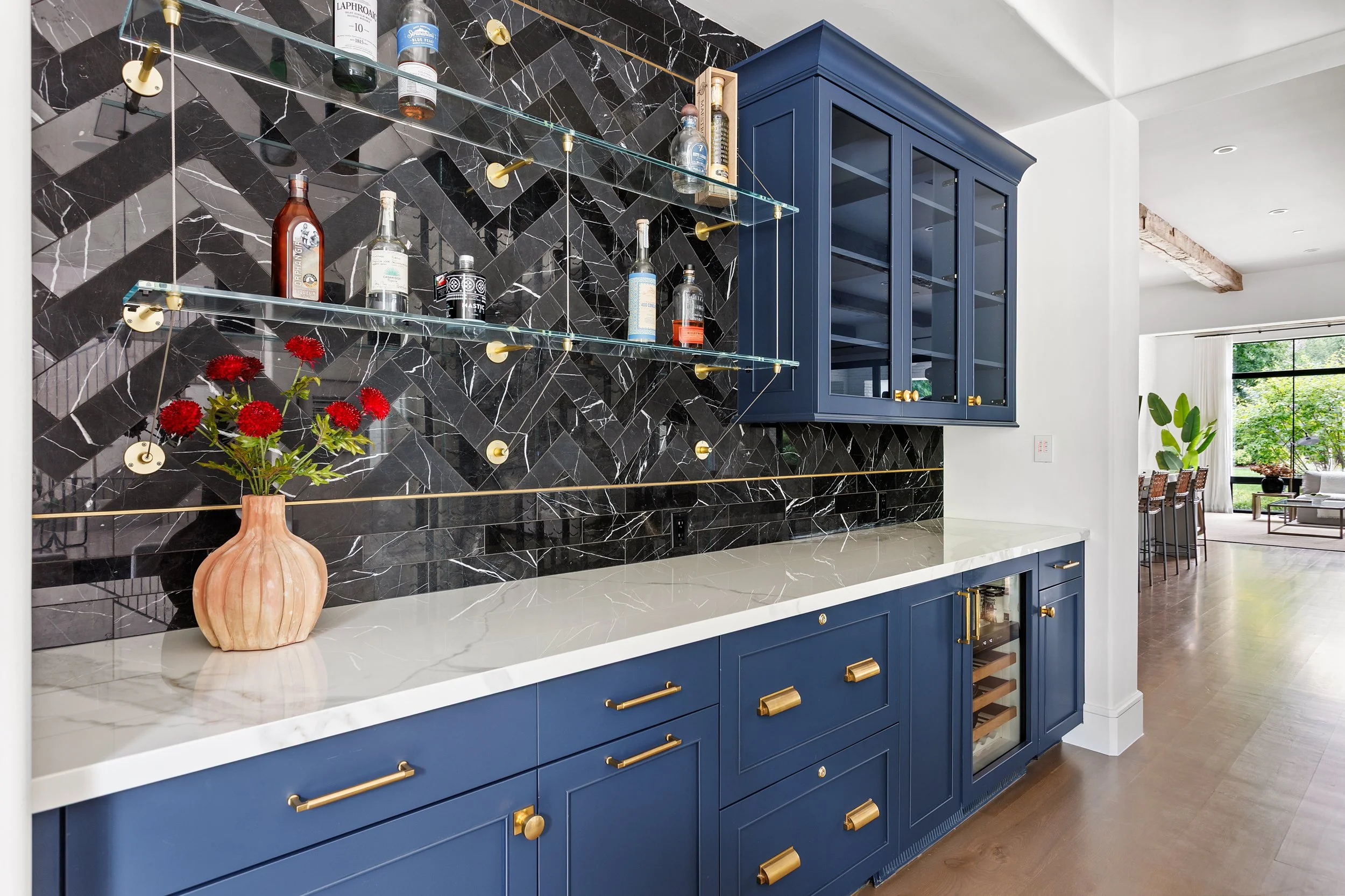 Modern kitchen with navy blue cabinets, white marble countertop, black herringbone marble backsplash, and glass shelves holding bottles. A beige vase with red flowers is on the countertop. Bright, open dining area with large windows and greenery outs
