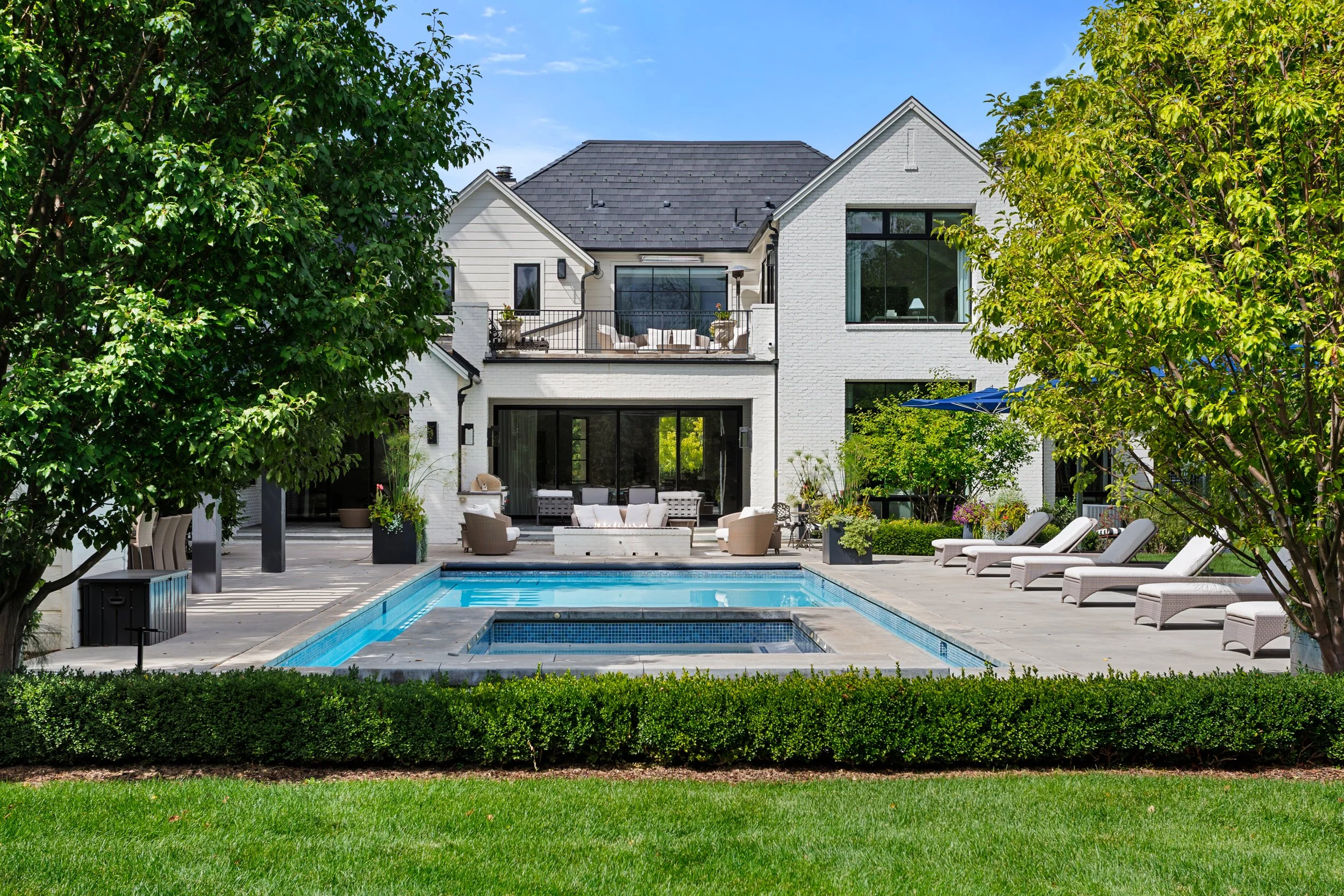 A backyard with a swimming pool, outdoor seating area, and a large white house with a balcony, surrounded by trees and greenery on a sunny day.
