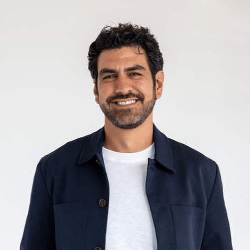 A smiling man with dark hair and a beard, wearing a white t-shirt and a dark jacket, standing against a plain background.