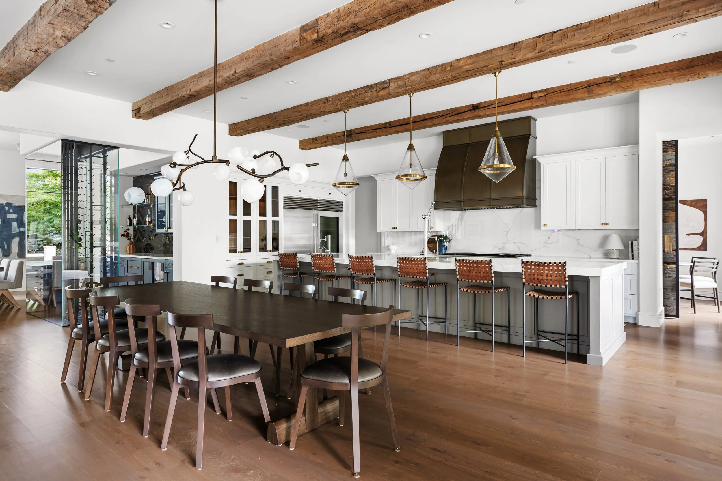Modern open kitchen and dining area with exposed wooden beams, white cabinets, marble backsplash, and hanging light fixtures.