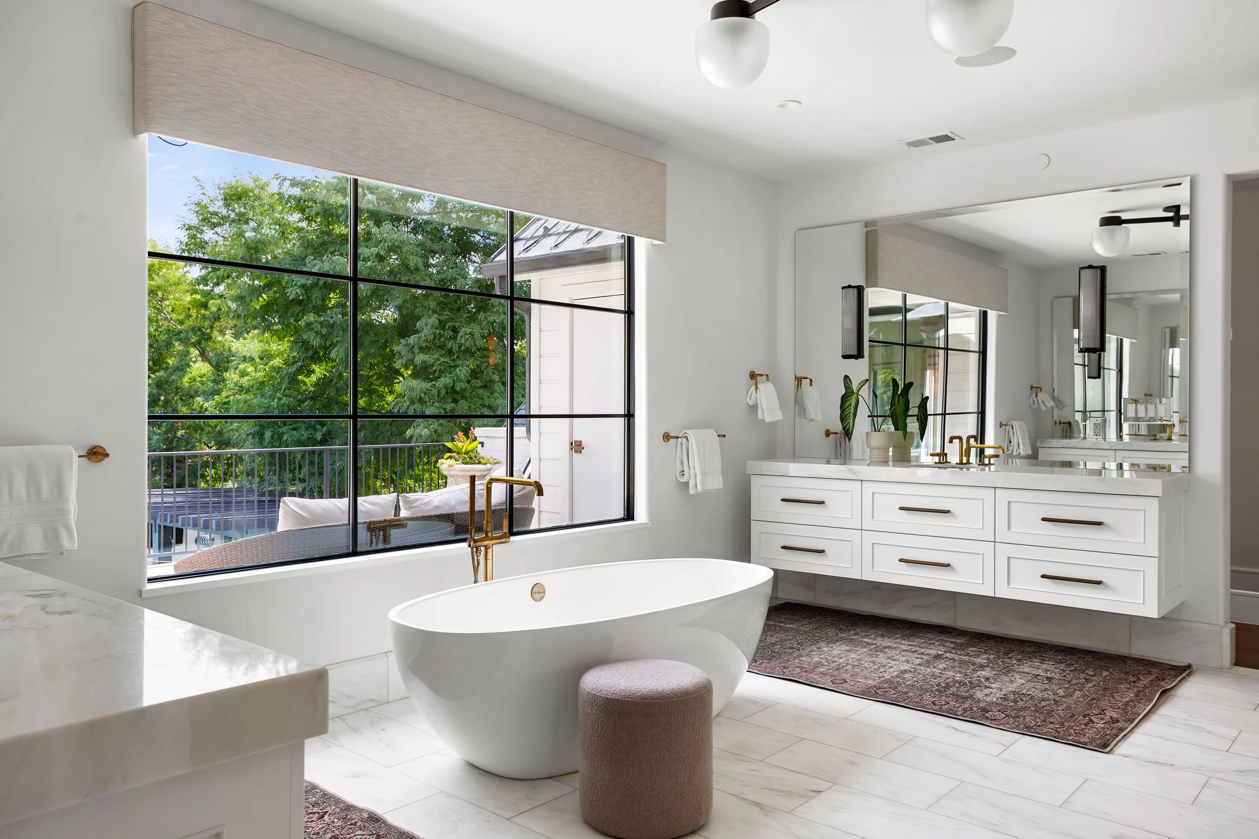 A bright bathroom with large windows showing green trees outside. It features a white freestanding bathtub, a vanity with a large mirror, gold fixtures, and towel racks. There is a potted plant on the vanity and a small stool near the tub. The floor 
