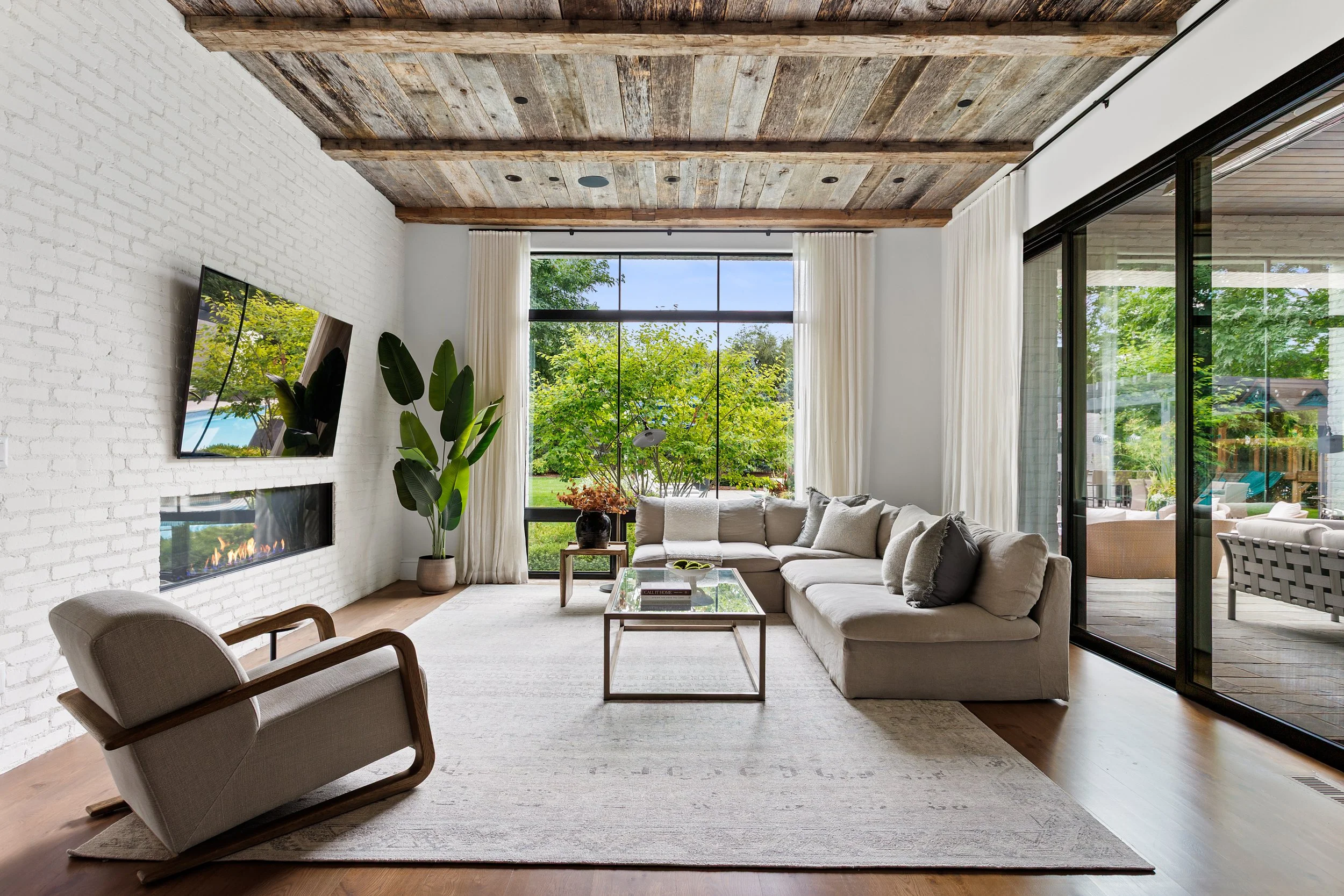 Living room with white brick fireplace, mounted TV, large window with curtains, beige sectional sofa, potted plant, glass coffee table, wood ceiling beams, and sliding glass door leading to outdoor patio with trees.