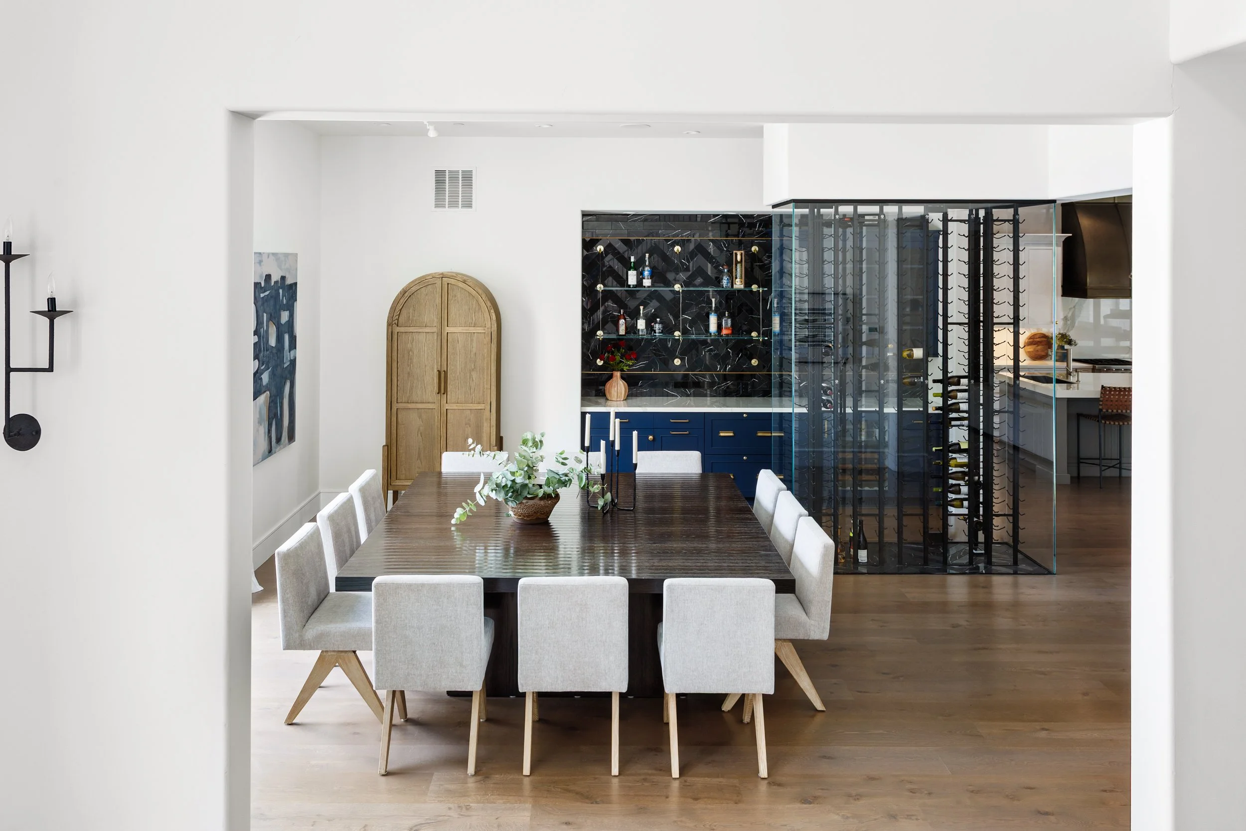 A dining room with a wooden table, surrounded by eight light-colored chairs, and a decorative centerpiece of flowers. In the background, there is a black and gold bar cabinet with bottles and glasses, a wooden arched door, art on the left wall, and a