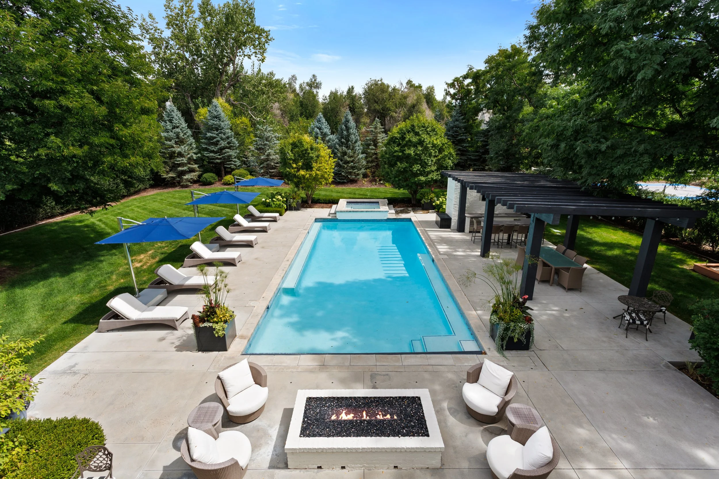 A backyard with a rectangular swimming pool surrounded by lounge chairs, umbrellas, and lush green trees.