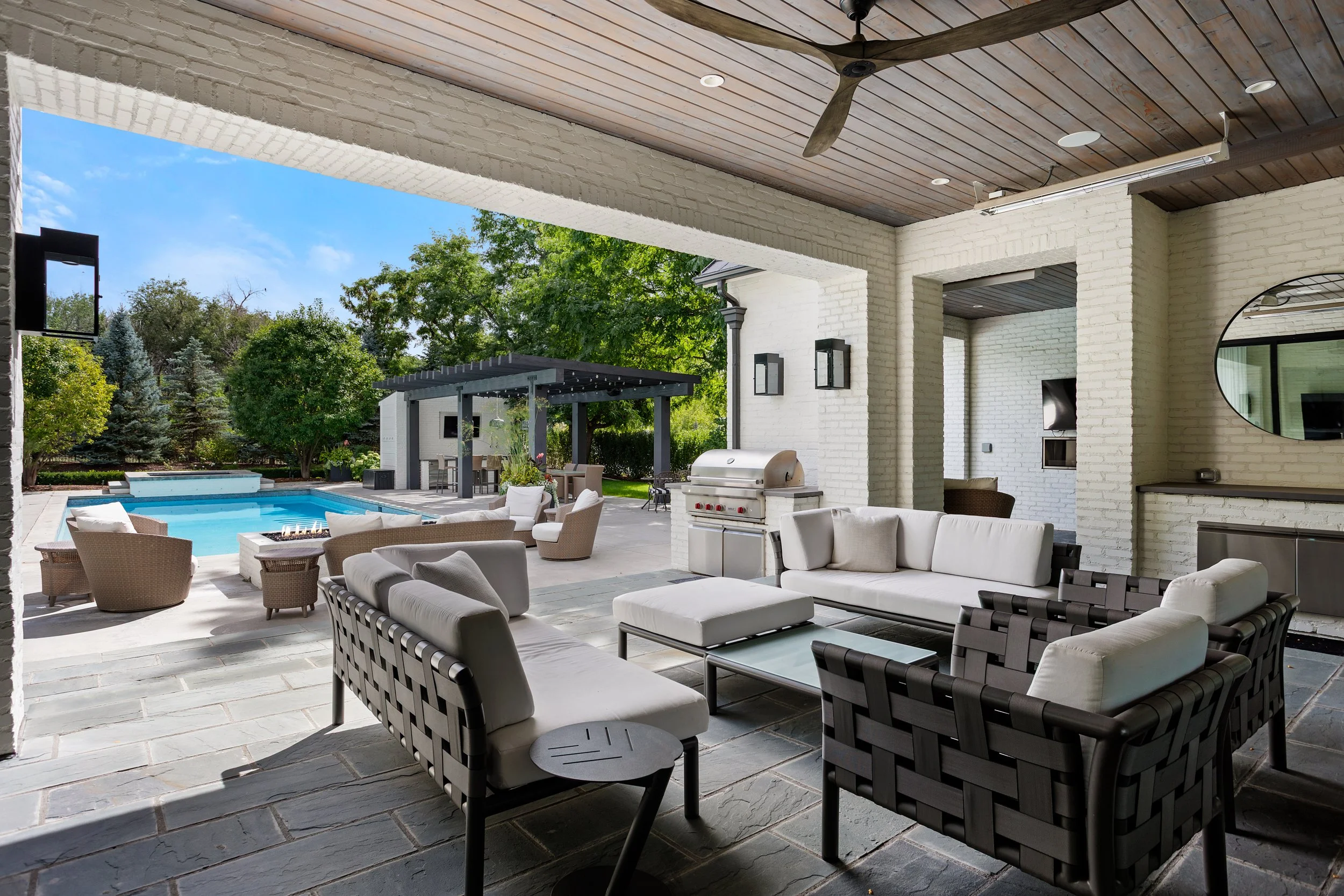 A covered outdoor patio area with seating, a grill, and a view of a swimming pool and garden with trees under a blue sky.