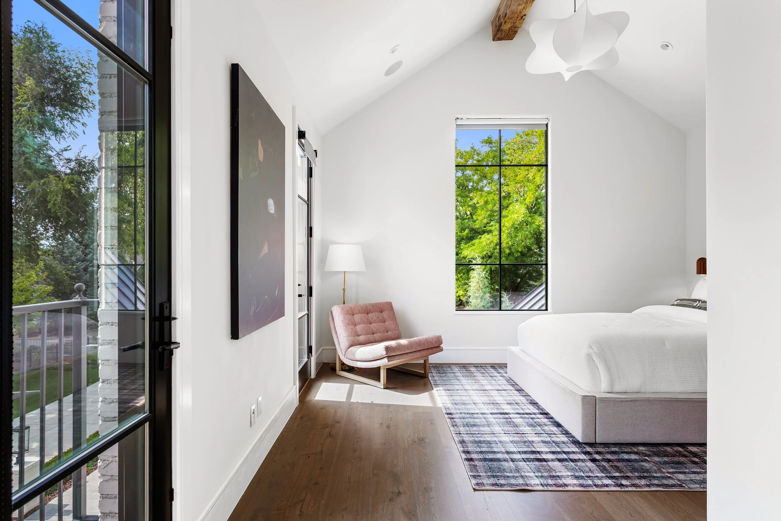 Bright modern bedroom with large window showing green trees outside, white walls, wooden floor, pink chair, white bed, black-framed artwork, and ceiling light fixture.