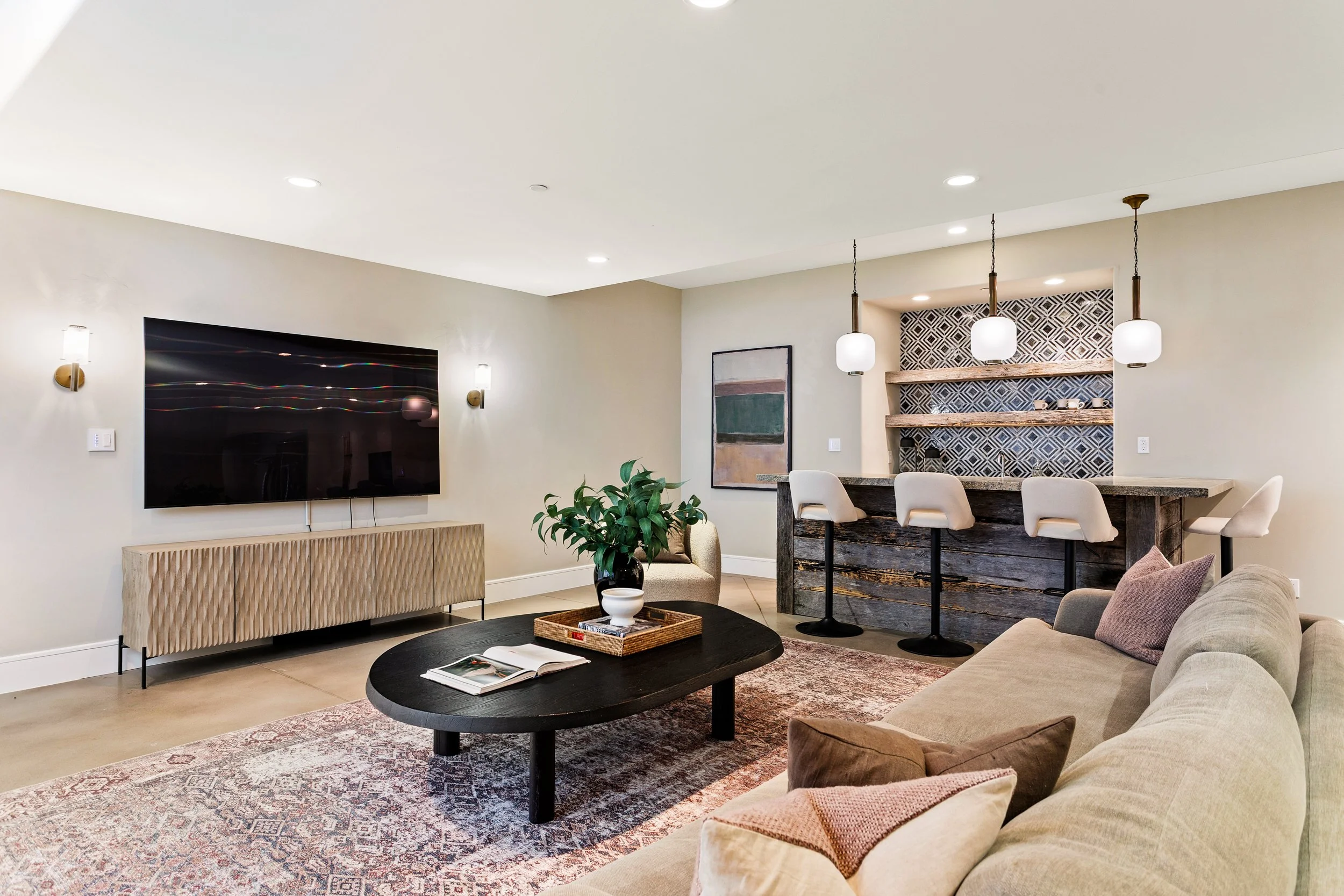 Living room with a wall-mounted television, beige furniture, a black coffee table with magazines, and a taupe couch with pillows. A wooden shelf with decorative items is above a bar area with white stools. Modern lighting fixtures hang from the ceili