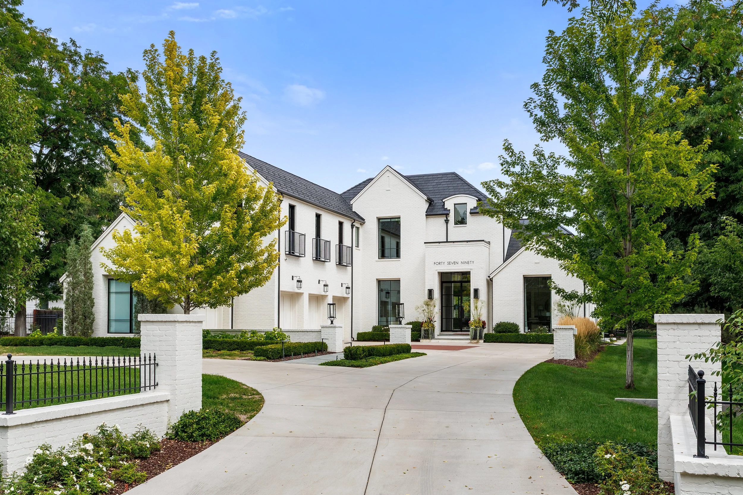 White modern house with black roof, surrounded by green trees and a landscaped front yard, with a curved sidewalk leading to the front entrance.