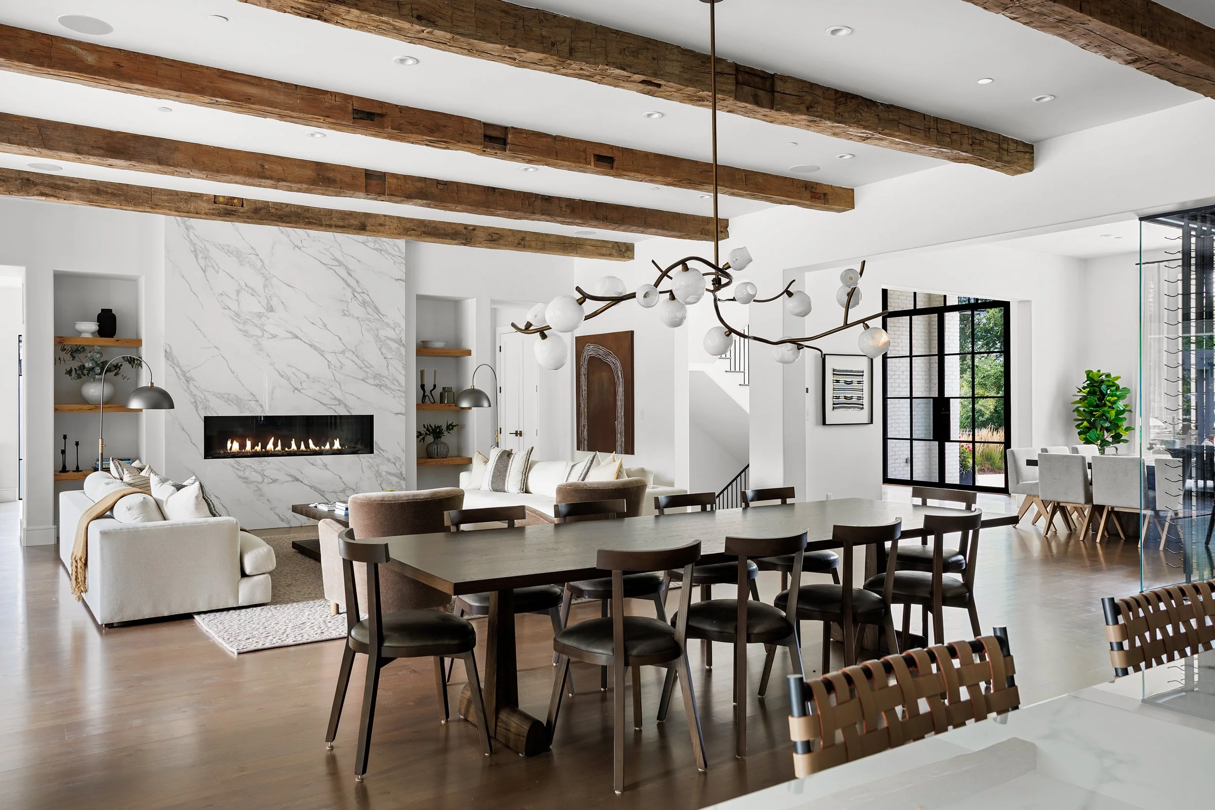 Interior of a modern open-concept living and dining area featuring a white sofa, a fireplace with a marble surround, a wooden dining table with black chairs, and a unique chandelier with globe-shaped lights, exposed wooden ceiling beams, and large fl