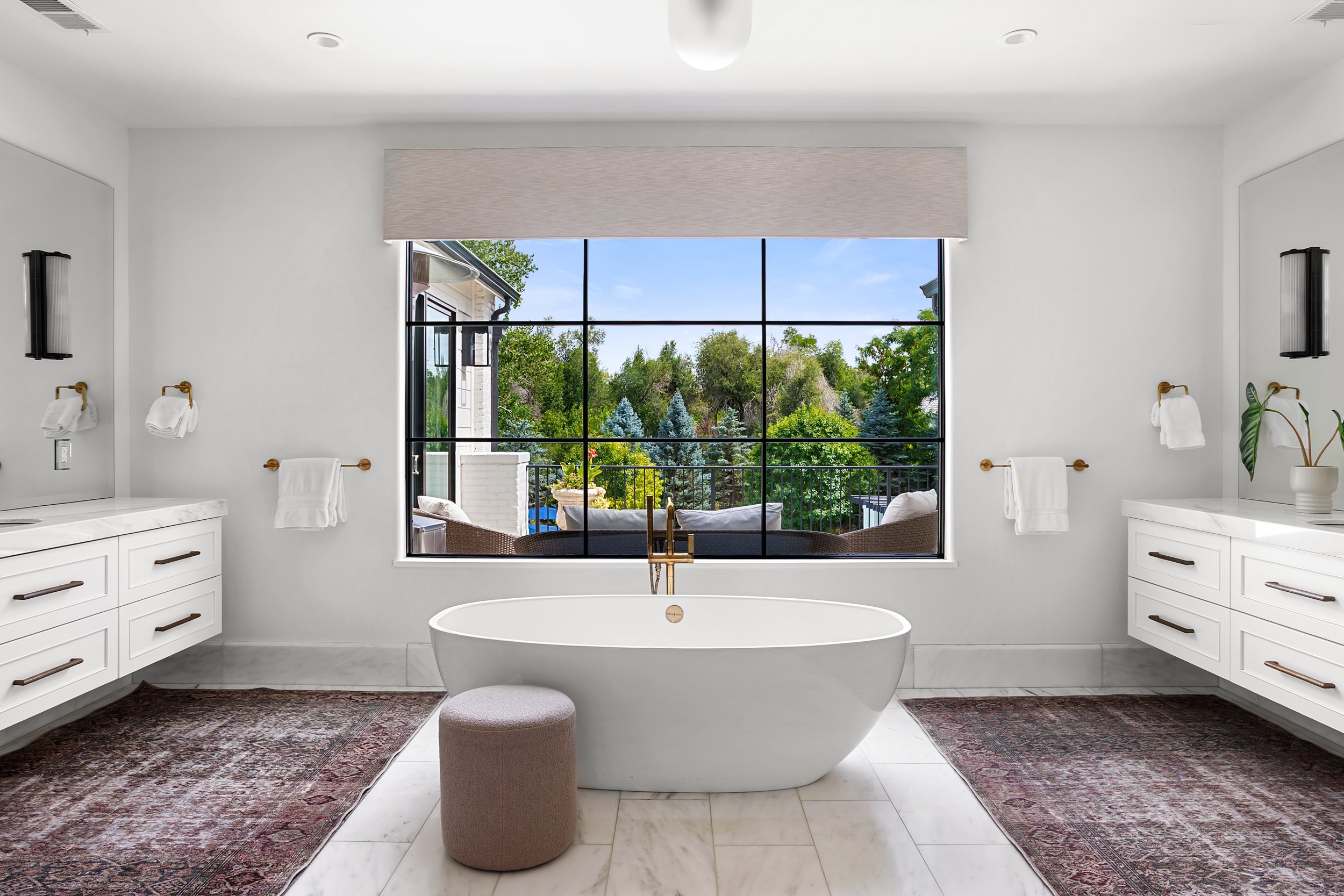 A bathroom with a large window overlooking a green outdoor area, a white freestanding bathtub, two vanities with white cabinetry, and decorative rugs on the marble floor.