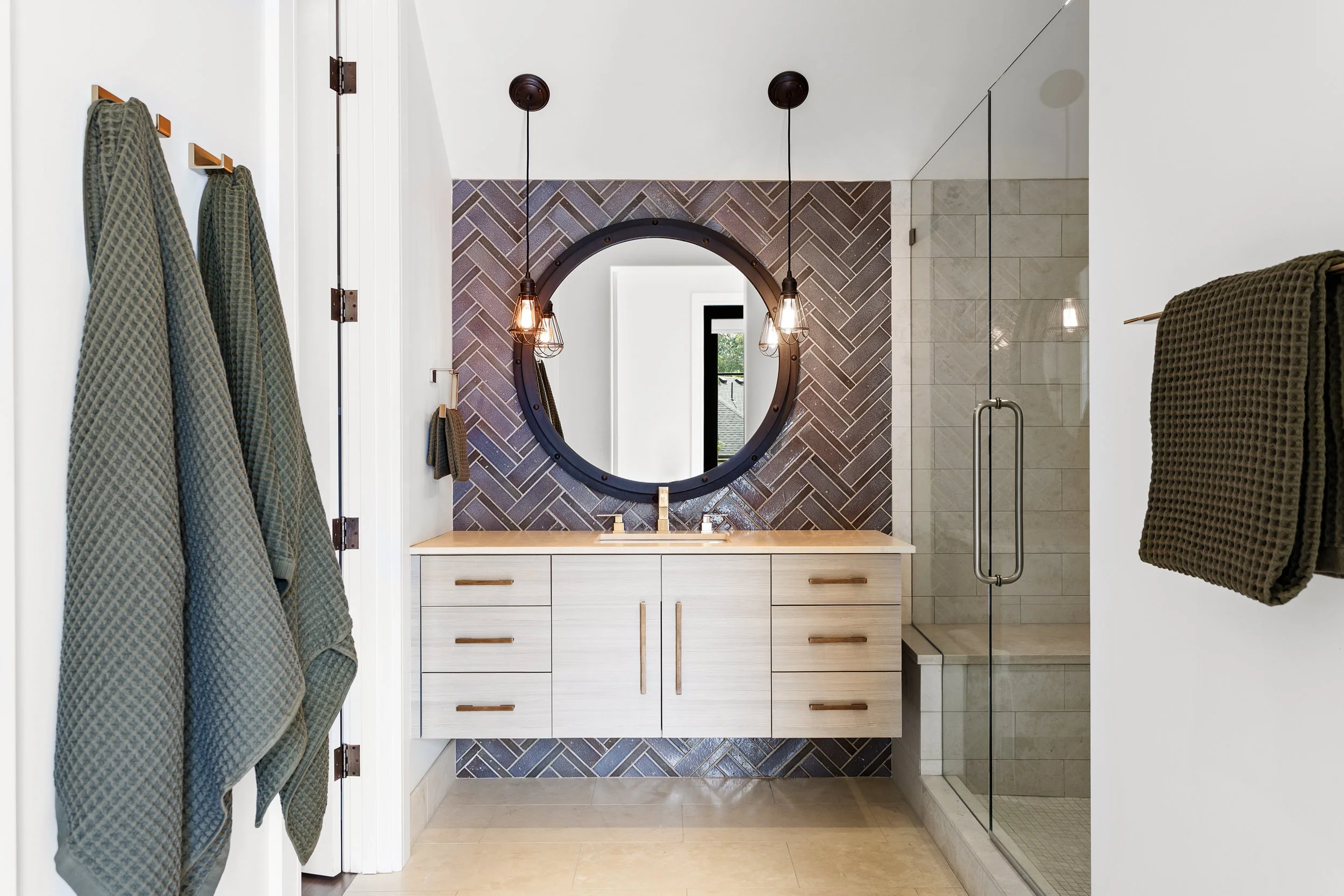 Modern bathroom with double vanity featuring a round mirror, pendant lights, towel hooks, and a walk-in shower with glass door.