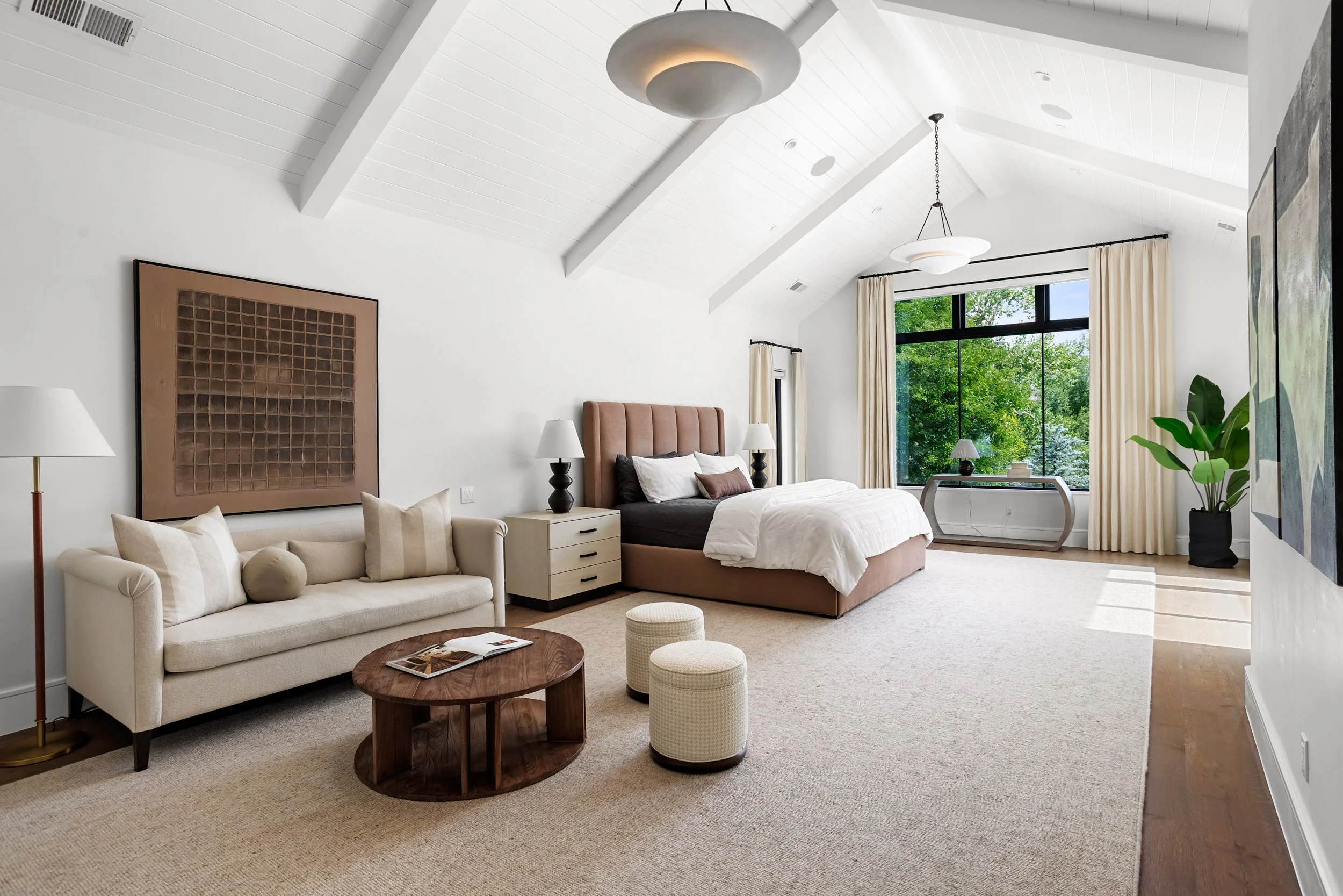 A spacious modern bedroom with a vaulted white ceiling and large window showing green trees outside. The room features a beige sofa with pillows, a wooden coffee table, a bed with a tan headboard, nightstands with lamps, and wall art. Light curtains 